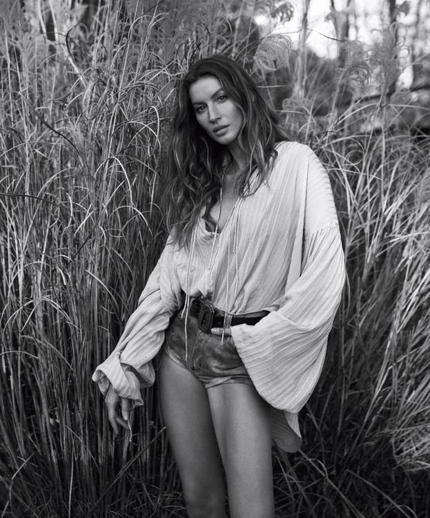 A woman with long hair standing among tall grass in a black and white photo, wearing a loose blouse and shorts.