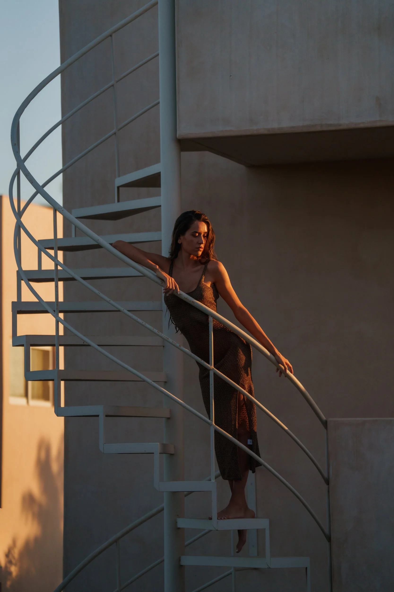 A woman wearing a black dress is standing on an outdoor spiral staircase, leaning against the handrail at sunset.