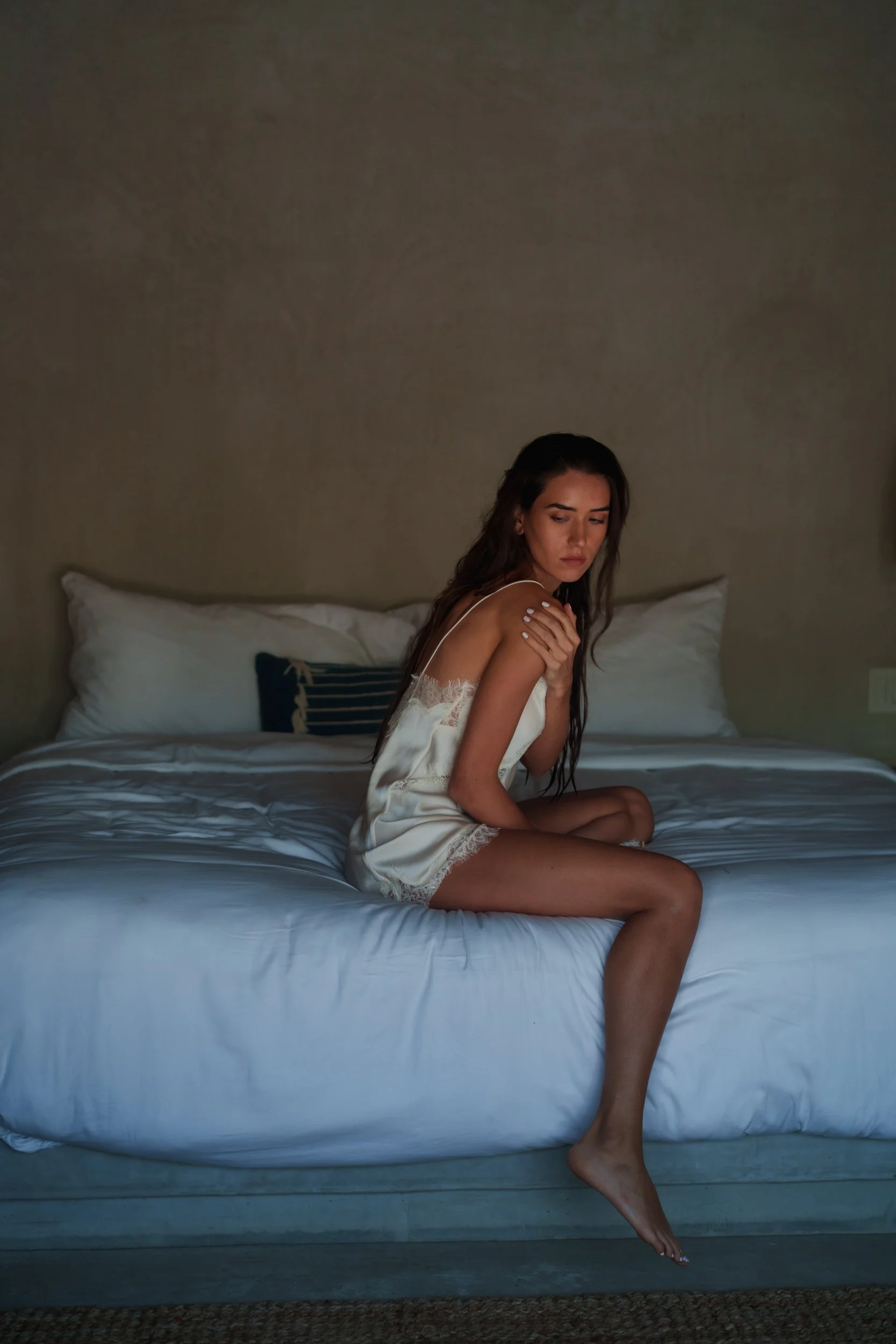 A young woman with long dark hair sitting on a bed in a dimly lit room, wearing a white slip dress with lace trim, looking down thoughtfully, with a neutral colored wall and pillows in the background.