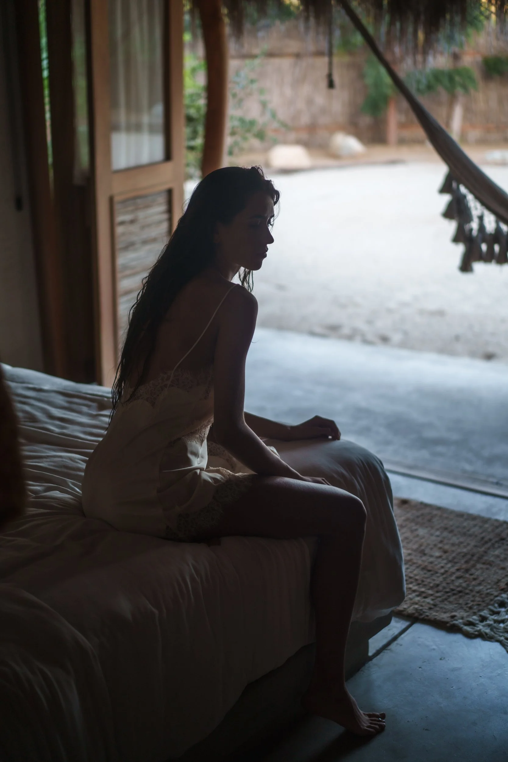 A woman with long, dark hair, wearing a light-colored sleepwear, sitting on her bed looking outside through an open door in a rustic, wooden house. Outside, there is a sandy yard with rocks and trees.