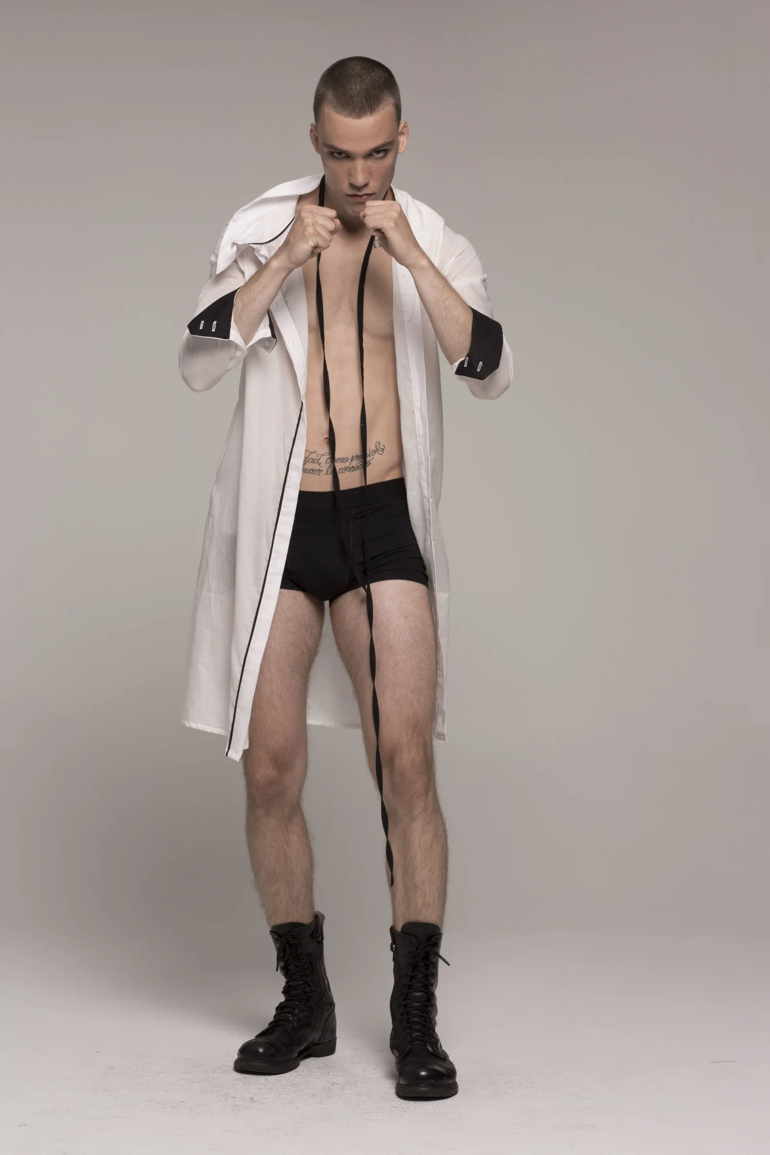 male model in boxing stance, and in sheer white collared dress with black stripe down center, black cuffs; dress unbuttoned, black shorts underneath