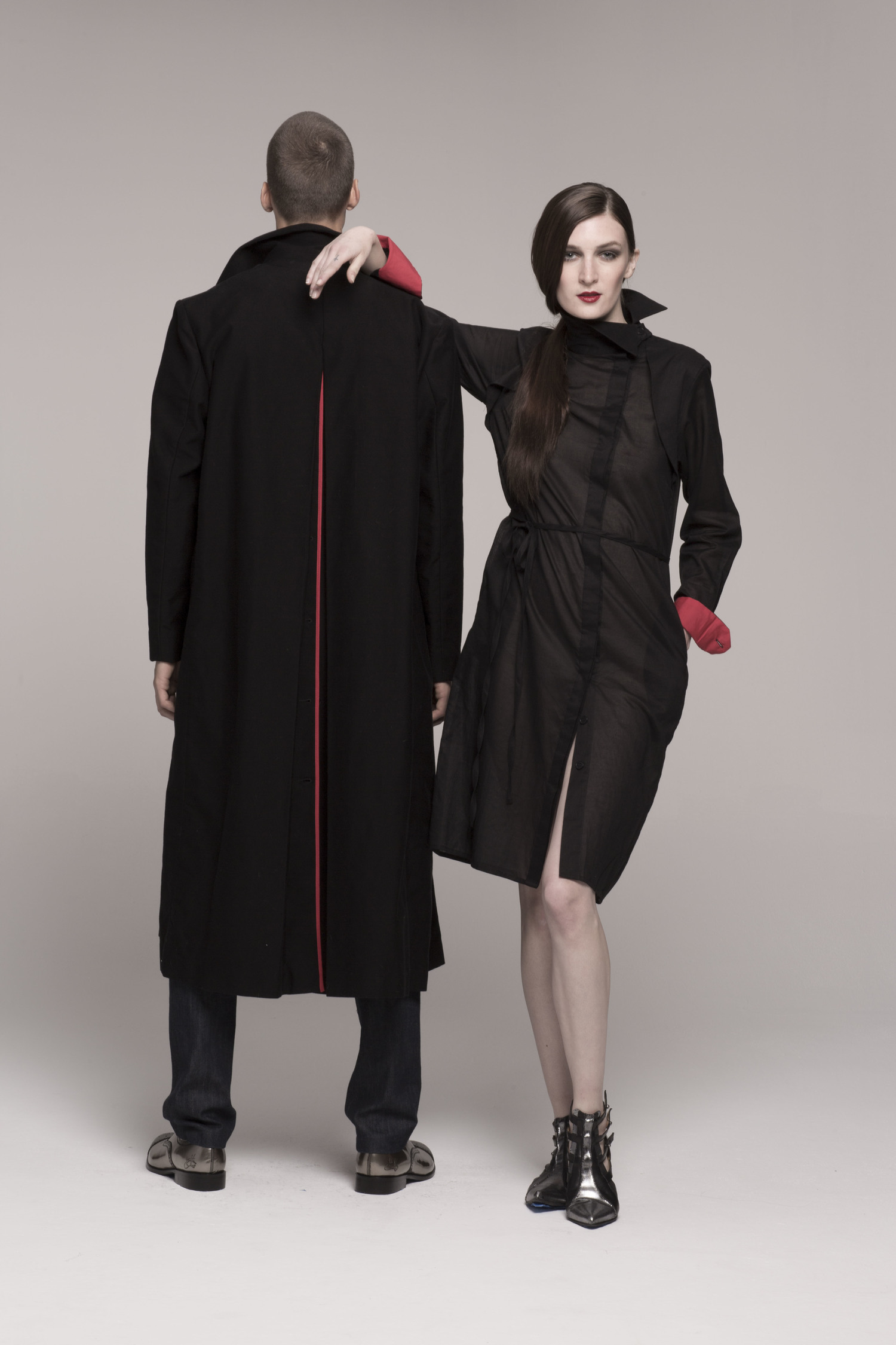 male model in long black coat, back to camera. Girl in sheer black dress with red cuffs, arm on model's shoulder