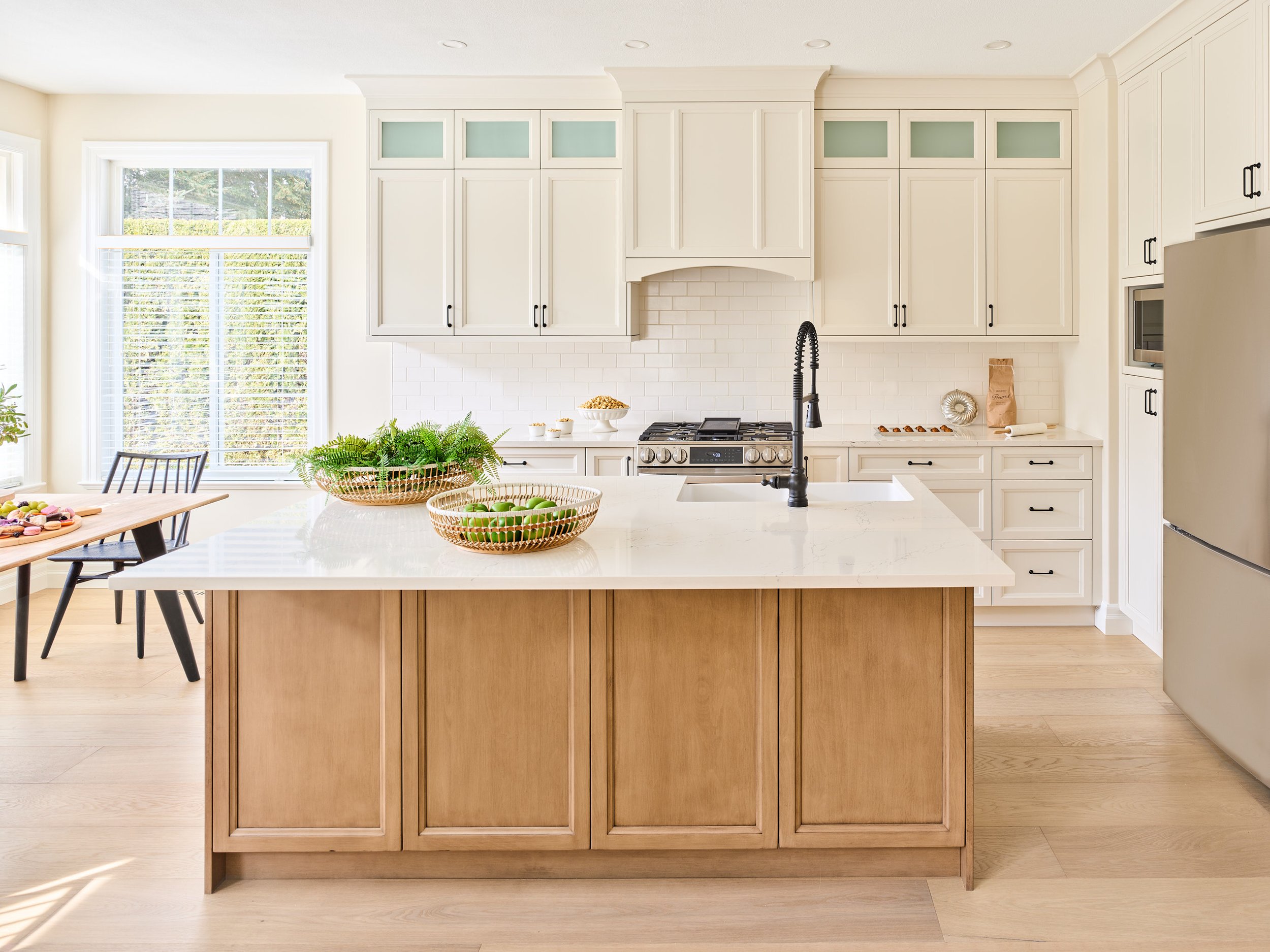 This sun-filled modern farmhouse kitchen features both painted and stained cabinetry with an island full of concealed storage. The range hood cover gracefully arches over creamy white subway tiles, stainless steel appliances, and wide plank white oa