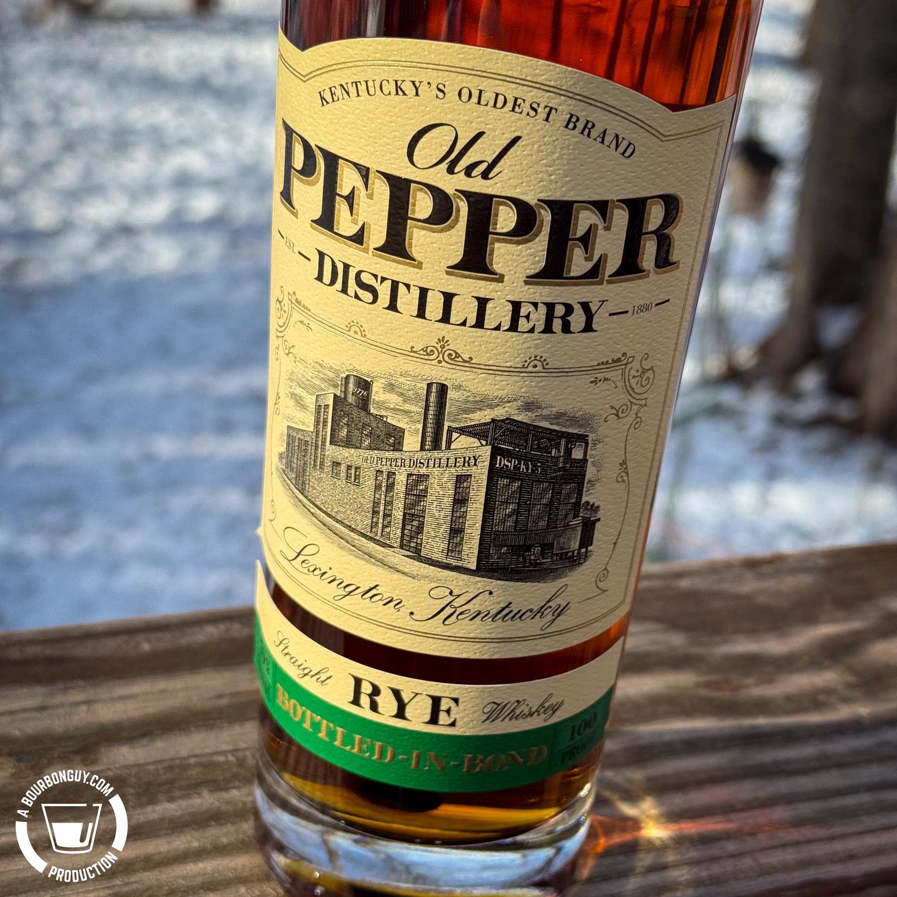 IMAGE: Close-up of an Old Pepper Distillery Bottled-in-Bond Straight Rye Whiskey bottle on a wooden railing, with snow-covered trees in the background and the vintage Lexington, Kentucky label in focus.