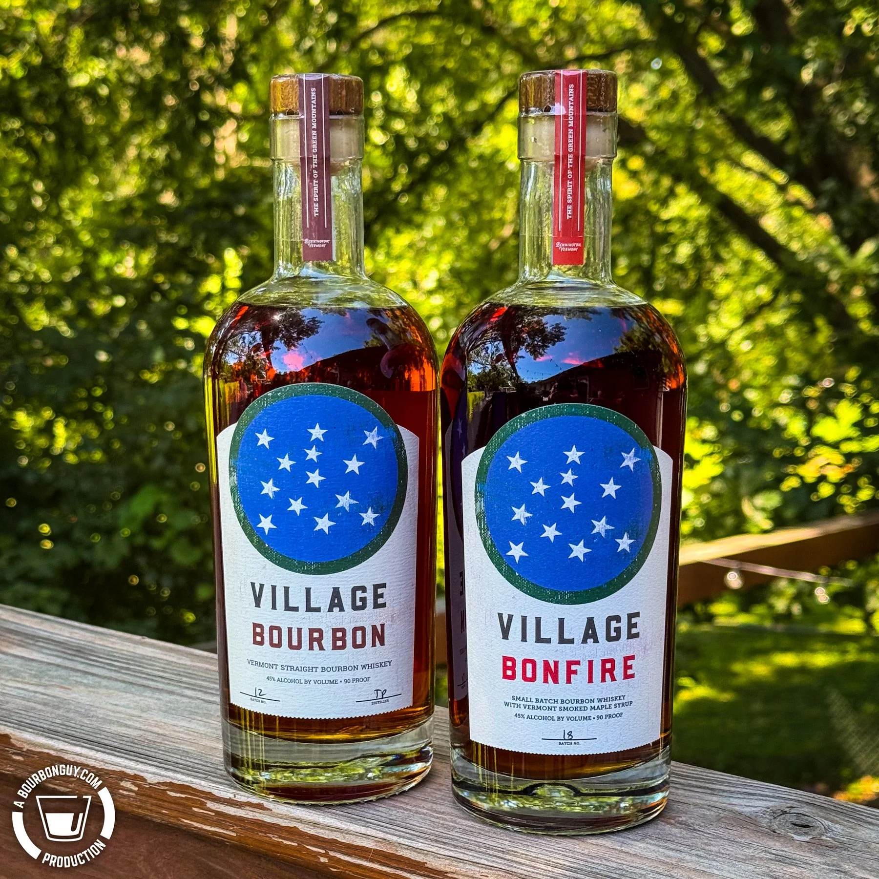 IMAGE: Two bottles of Village Garage Distillery spirits—Village Bourbon and Village Bonfire—standing side by side on a weathered wooden deck railing, with lush green trees softly blurred in the background on a sunny summer day.