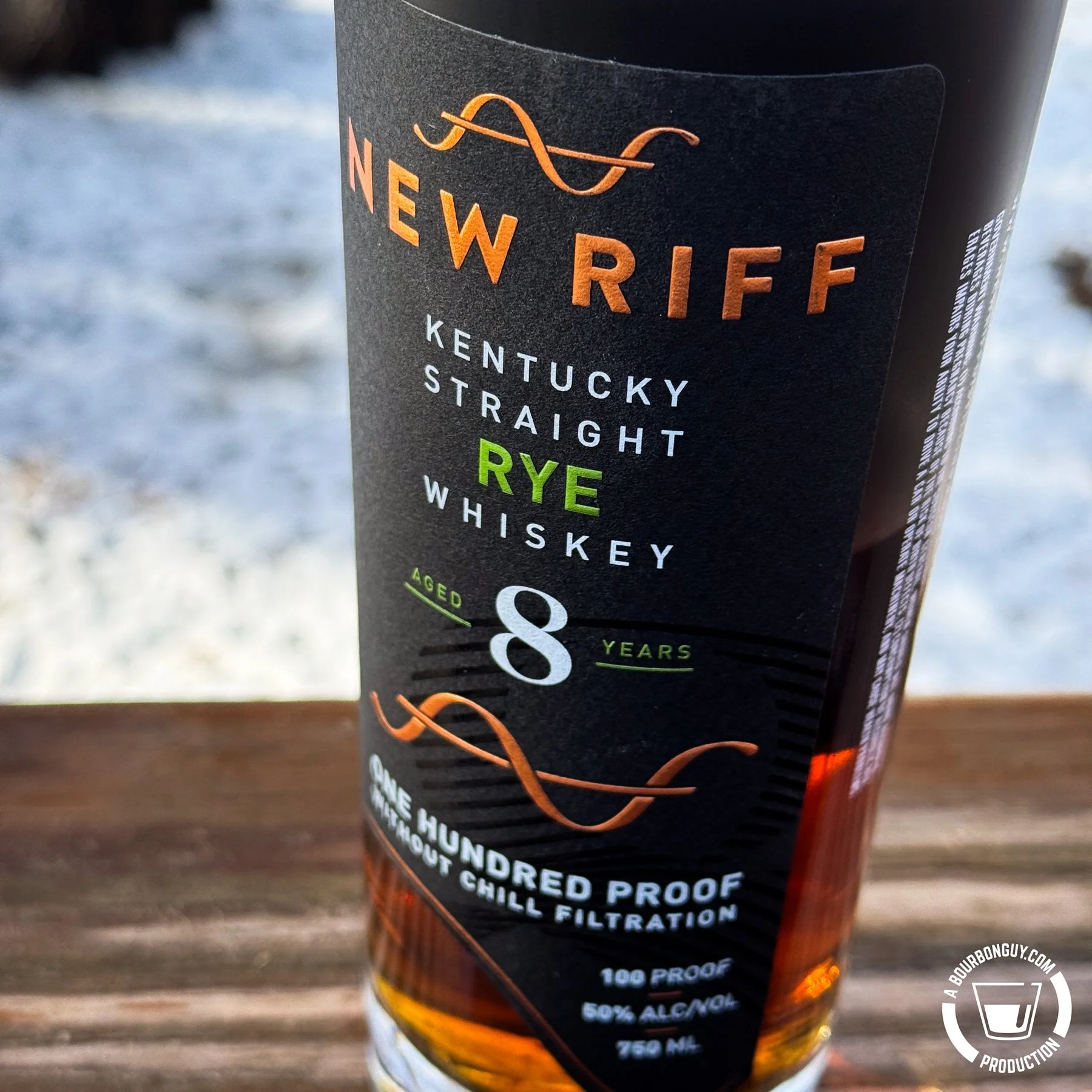 IMAGE: New Riff Kentucky Straight Rye Whiskey aged 8 years, 100 proof, photographed outdoors on a snowy wooden railing with amber whiskey glowing through the bottle.