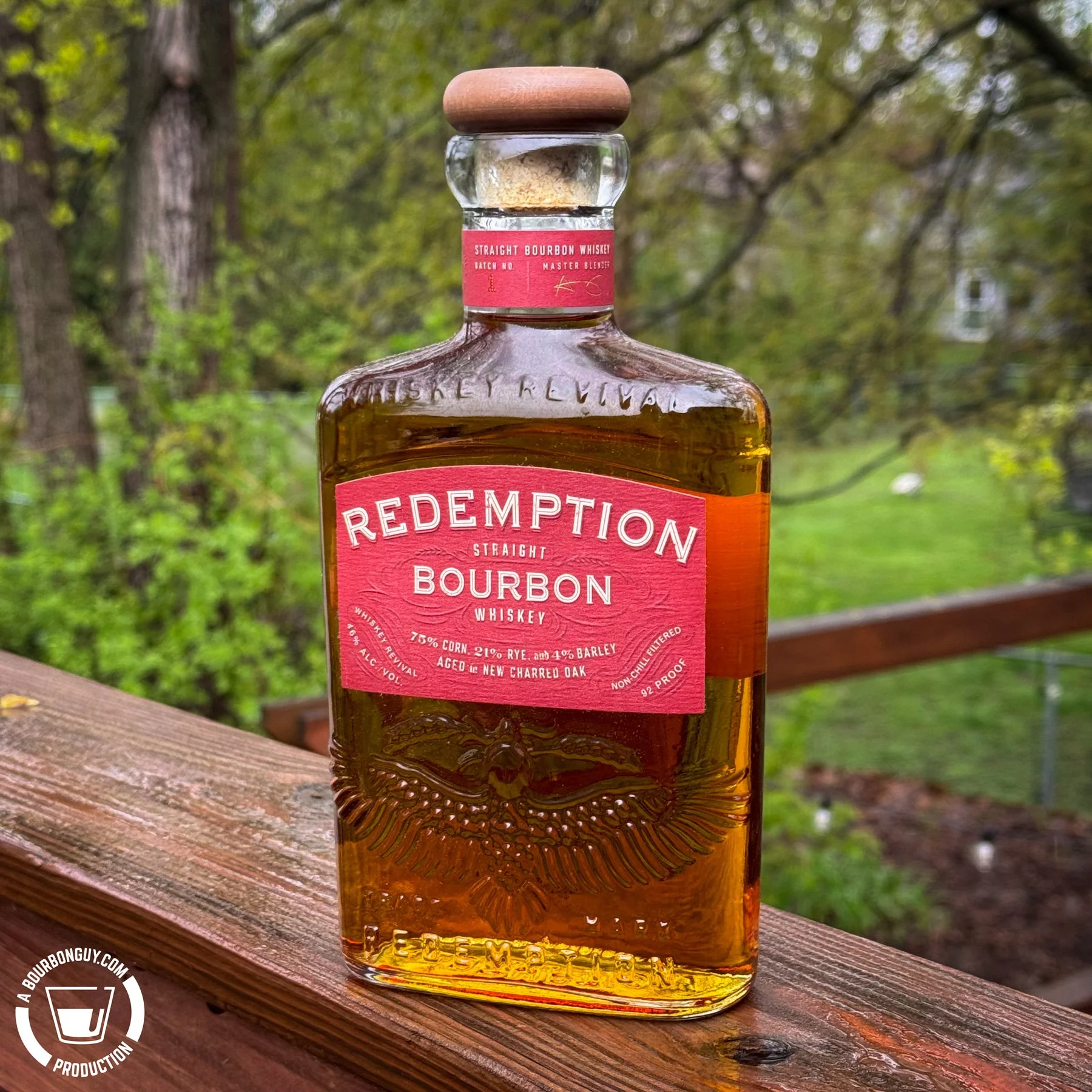 IMAGE: Redemption Bourbon bottle with red label and wood-topped cork sitting on a deck railing outdoors, green trees blurred in the background, BourbonGuy watermark in lower left.