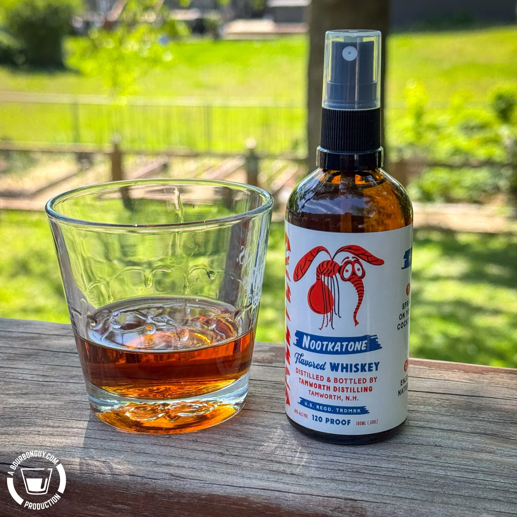 IMAGE: Glass with a whiskey cocktail beside a spray bottle of Tamworth Distilling Nootkatone flavored whiskey on a deck railing overlooking a sunny backyard.
