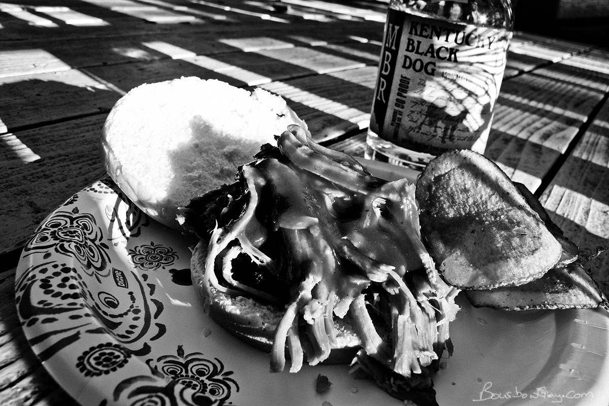 A pulled-pork sandwich with chips and a bottle of MB Roland Kentucky Black Dog in the background.