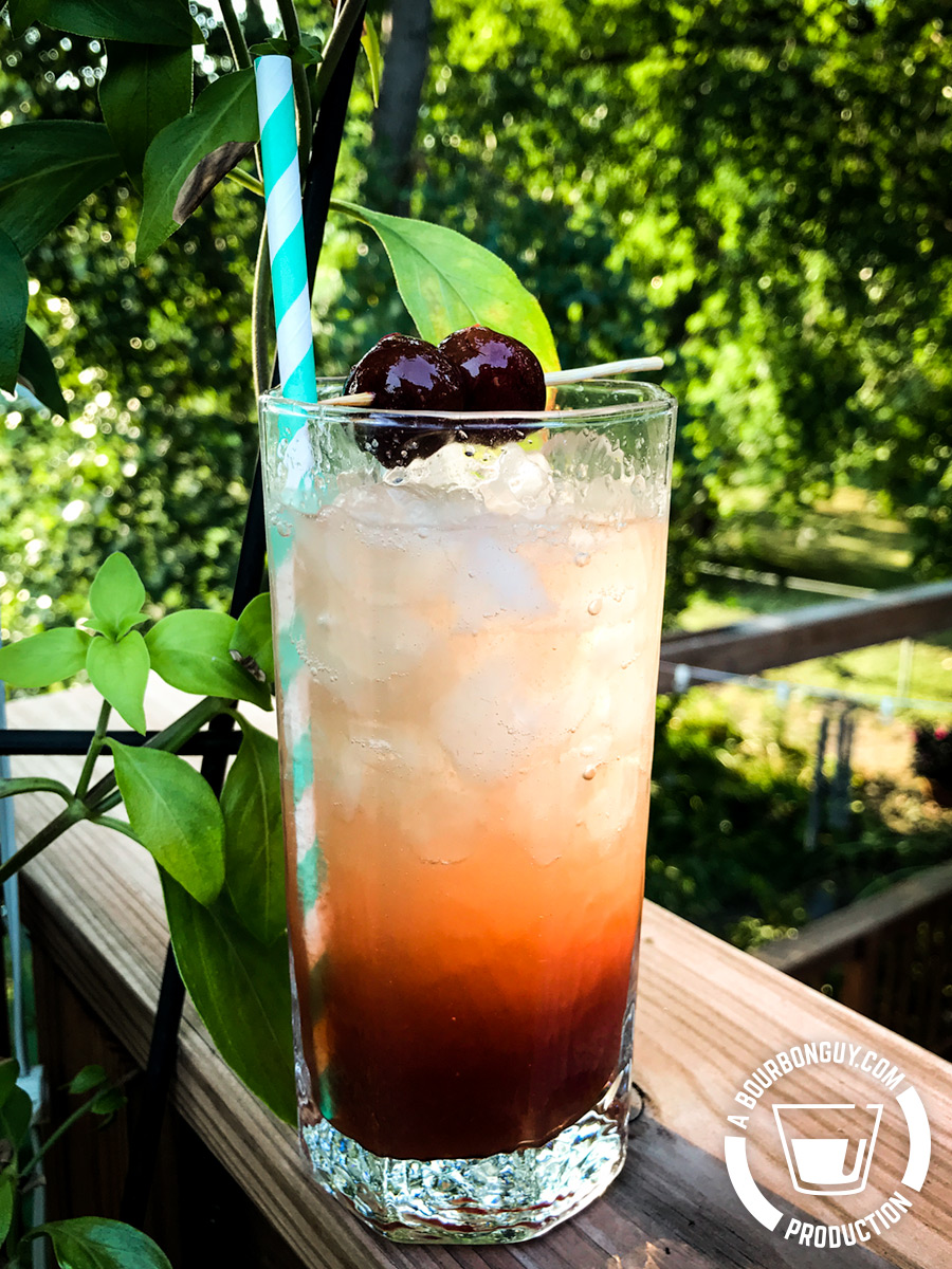IMAGE: An antique collins glass filled with liquid ranging from clear at the top to a cloudy red at the bottom. Two cherries on a skewer are balanced on the rim as a garnish.
