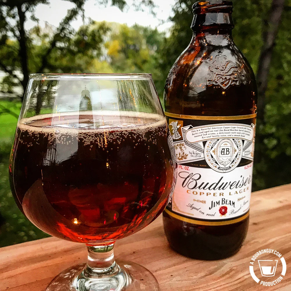 IMAGE: a glass of Budweiser Copper Lager. Rich copper color in a tulip style beer glass. Behind it is a bottle of the beer.
