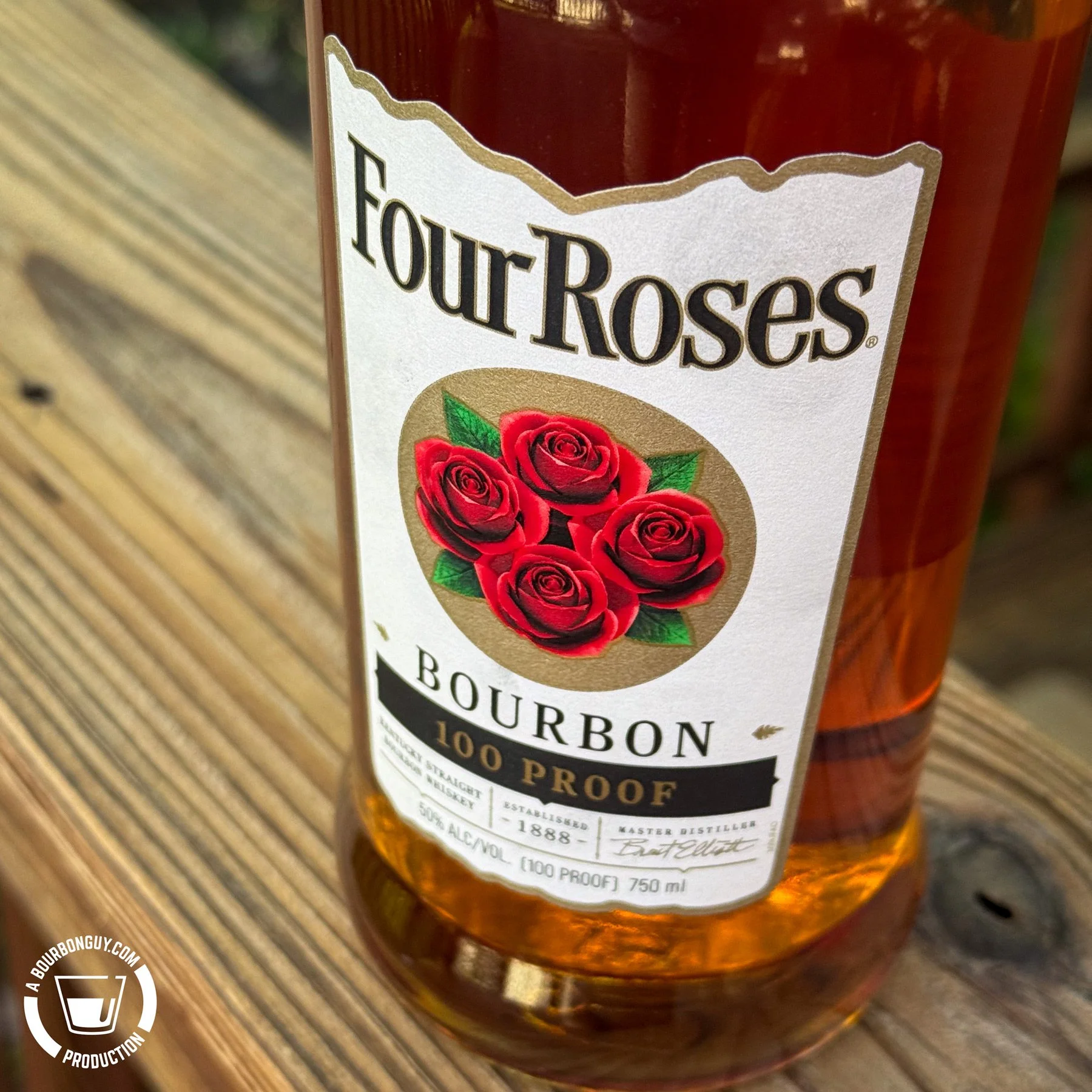 IMAGE: Close-up of a Four Roses Bourbon 100 Proof bottle on a wooden deck railing, showing its white label with the iconic four red roses.