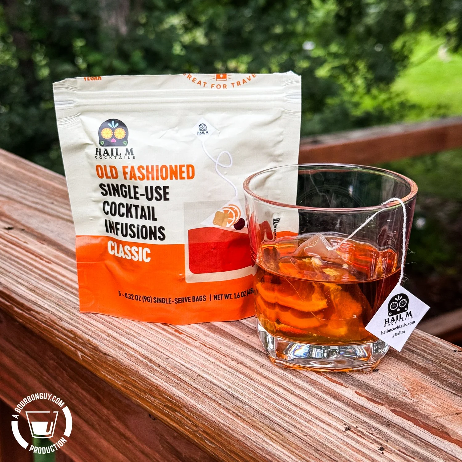 IMAGE: A bag of Hail M Cocktails Old Fashioned Single-Use Cocktail Infusions (Classic) with a glass of bourbon containing an infusion bag, sitting on a wooden deck railing with greenery in the background.