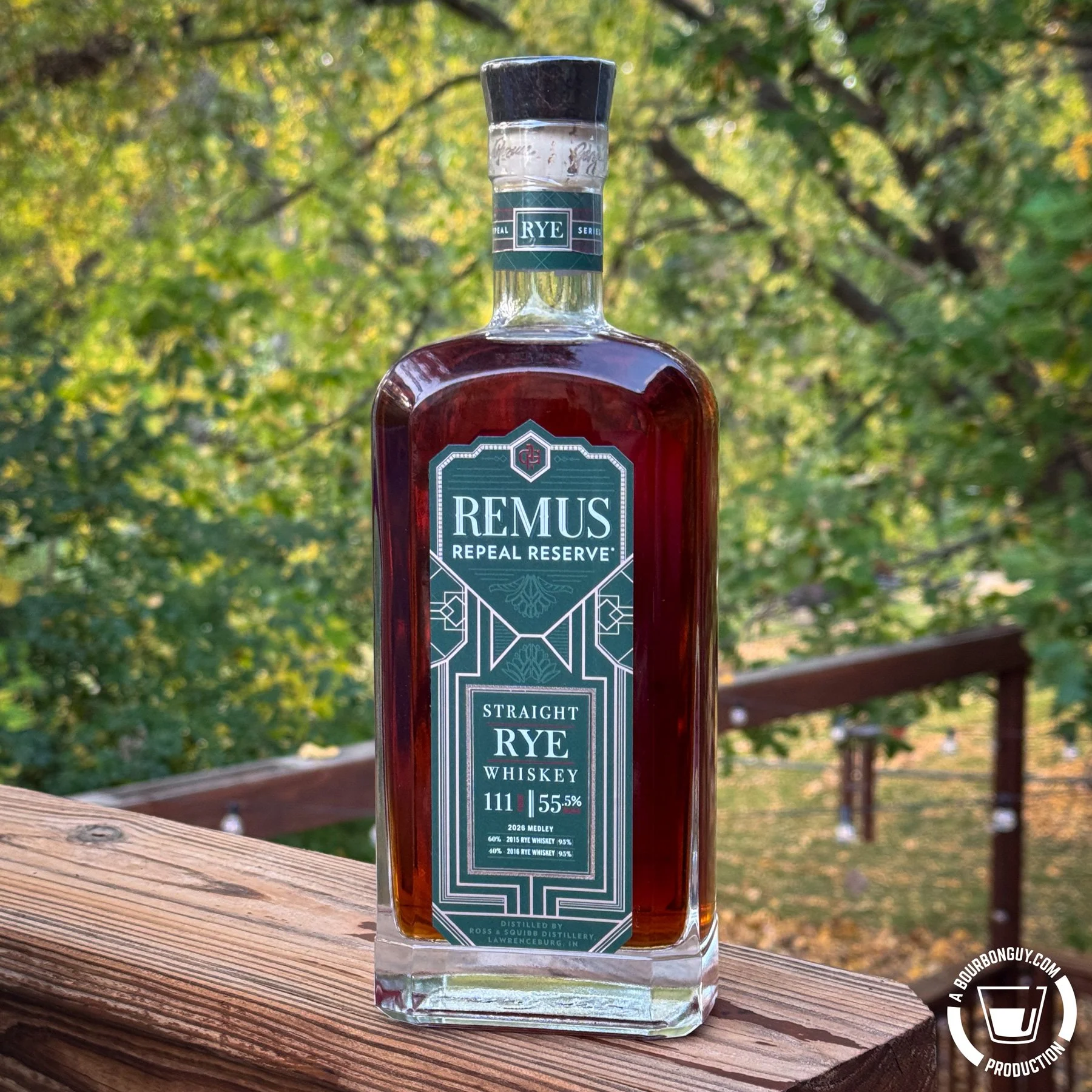 IMAGE: A bottle of Remus Repeal Reserve Straight Rye Whiskey, 111 proof, sits on a weathered wooden deck railing with green and yellow autumn foliage in the background.