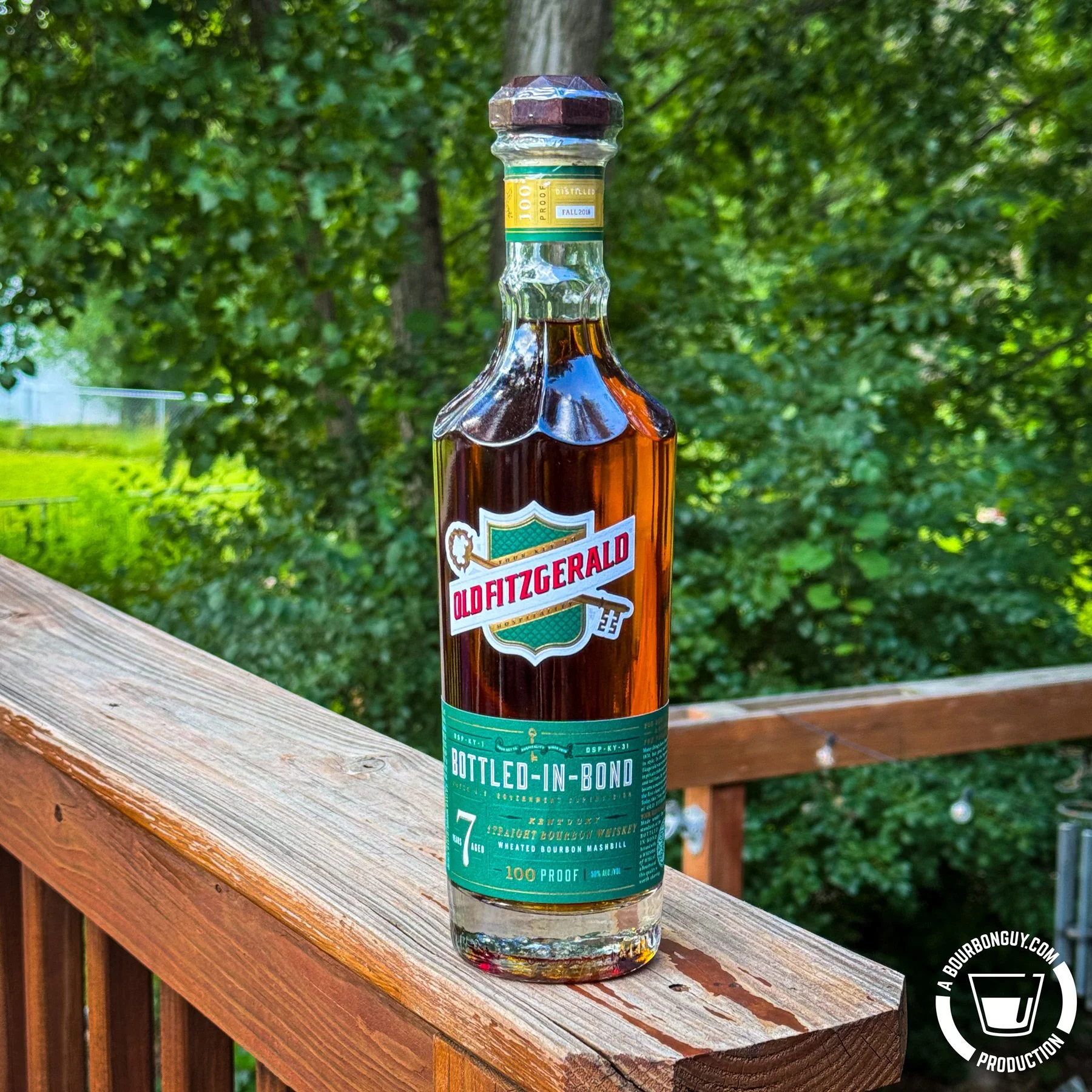 IMAGE: Bottle of Old Fitzgerald Bottled-in-Bond 7-Year-Old bourbon standing on a weathered wooden deck railing, with lush green trees softly blurred in the background on a sunny summer day.