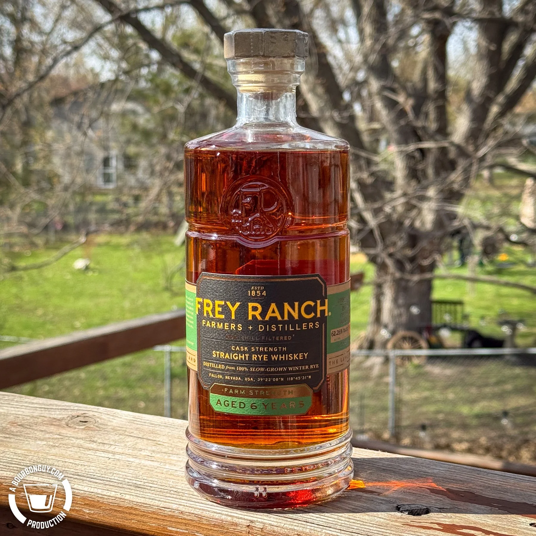 IMAGE: Frey Ranch Cask Strength Straight Rye Whiskey (aged 6 years) bottle on a wooden deck with backyard trees in the background, showing green and black label details and BourbonGuy watermark in the corner