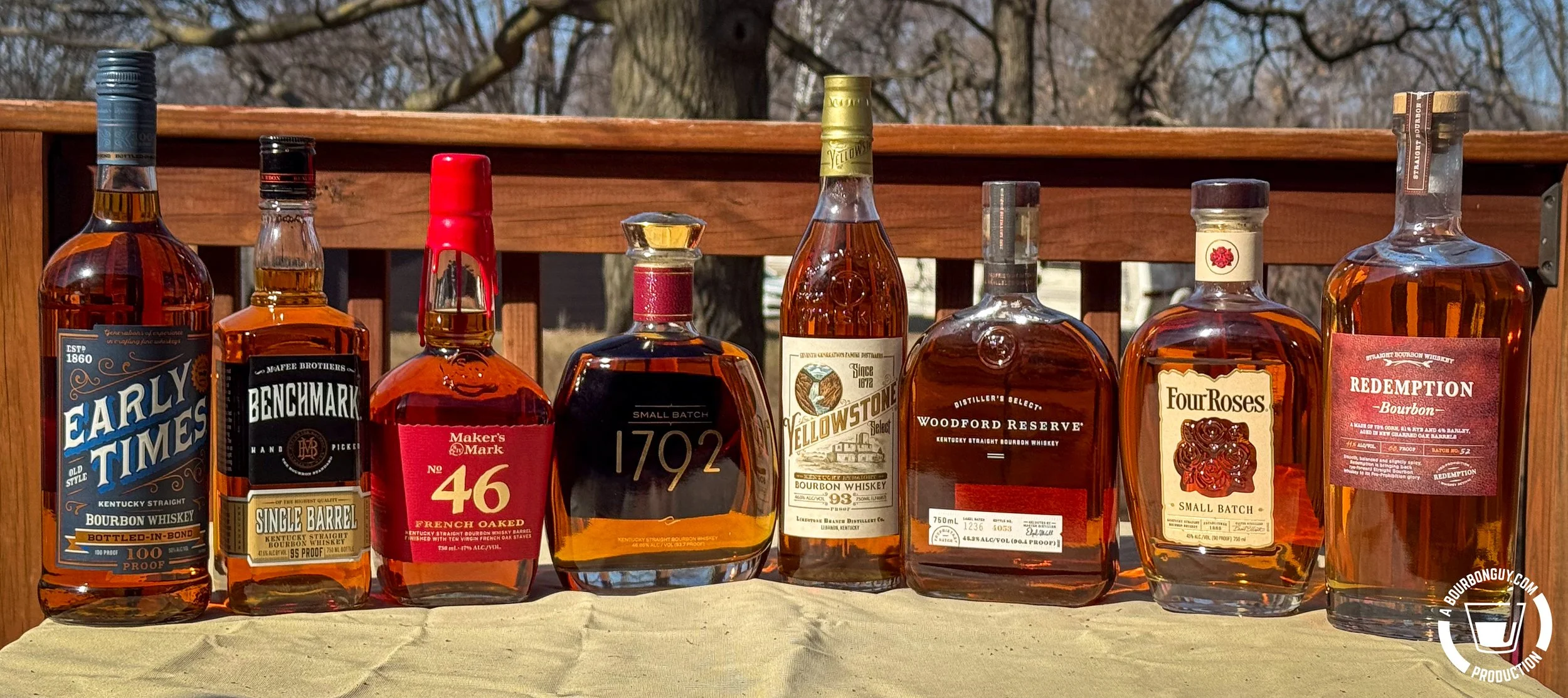 IMAGE: Eight bourbon bottles lined up on a deck, including Early Times, Benchmark, Maker’s 46, 1792, Yellowstone, Woodford Reserve, Four Roses, and Redemption, with trees in background
