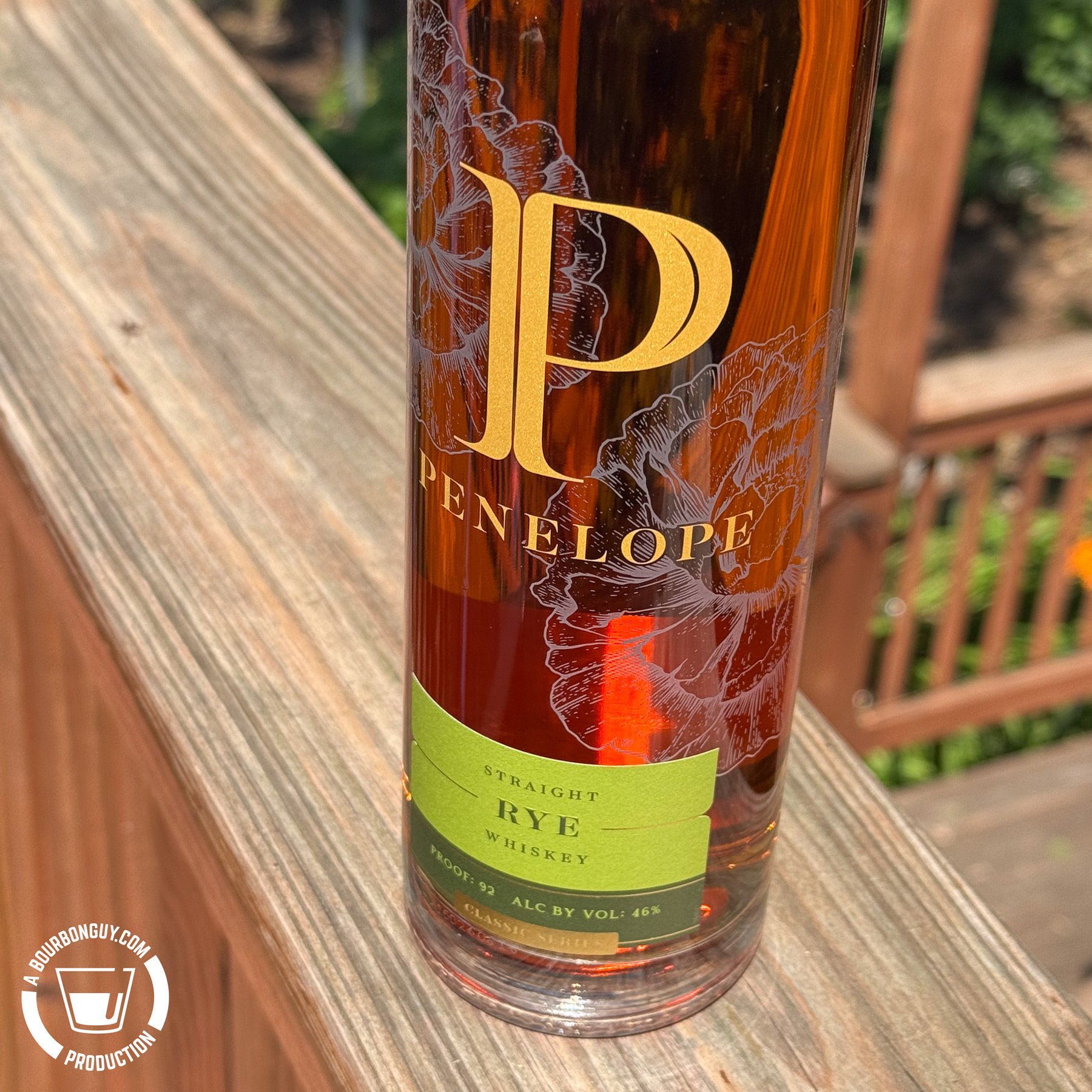 IMAGE: Close-up of a Penelope Straight Rye Whiskey bottle standing on a weathered wooden deck railing, with the gold logo and green label prominently displayed against a softly blurred backyard background on a sunny day.
