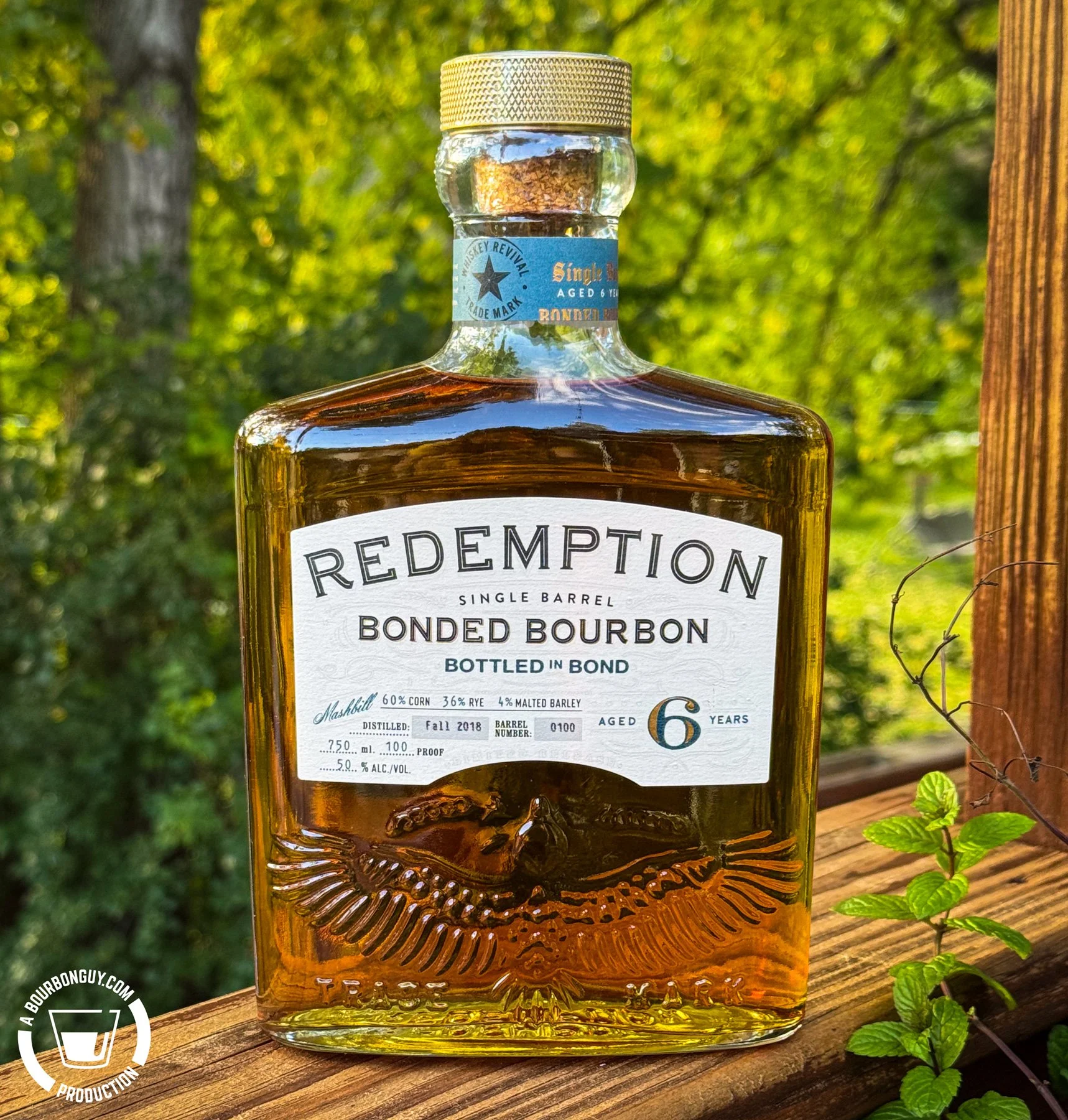 IMAGE: Bottle of Redemption Single Barrel Bonded Bourbon, aged 6 years and bottled at 100 proof, sitting on a wooden deck railing against a sunlit green background.