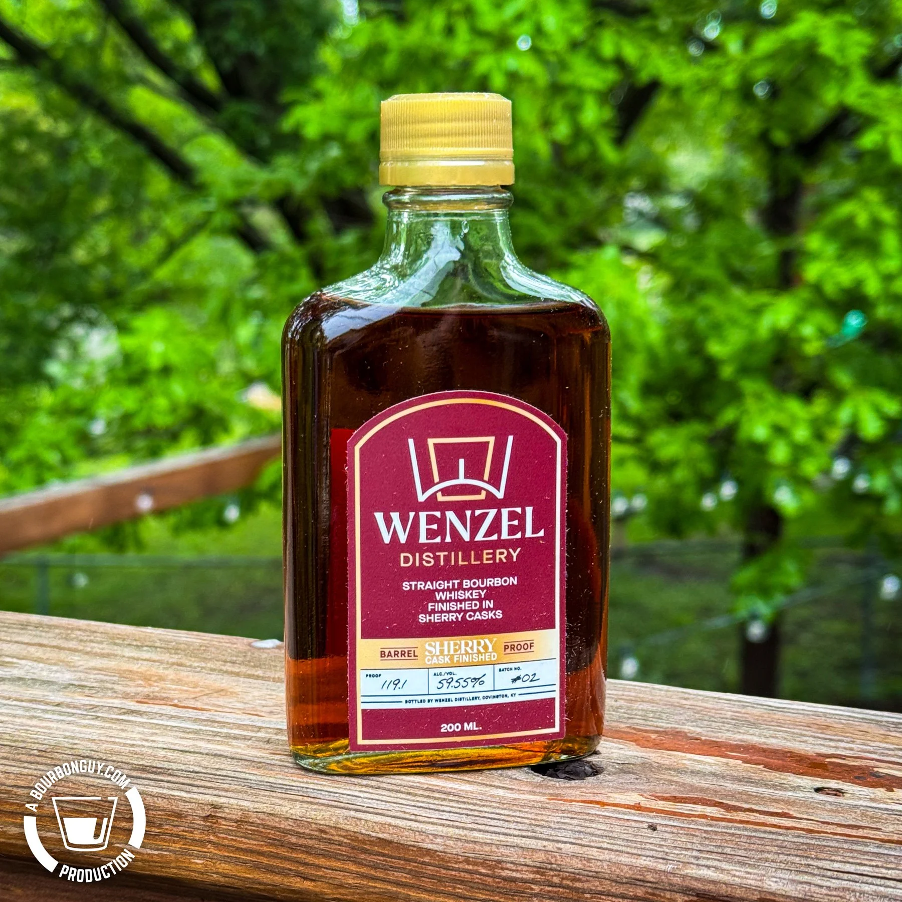 IMAGE: Small bottle of Wenzel Distillery Straight Bourbon Whiskey finished in sherry casks sitting on a deck railing with bright green trees blurred in the background.