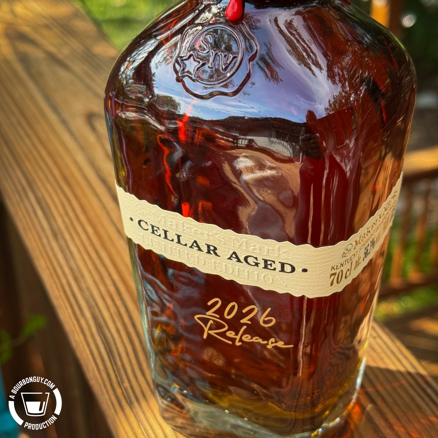 IMAGE: Close-up of a Maker’s Mark Cellar Aged 2026 Release bourbon bottle on a wooden deck railing, with amber whiskey glowing in the sunlight.