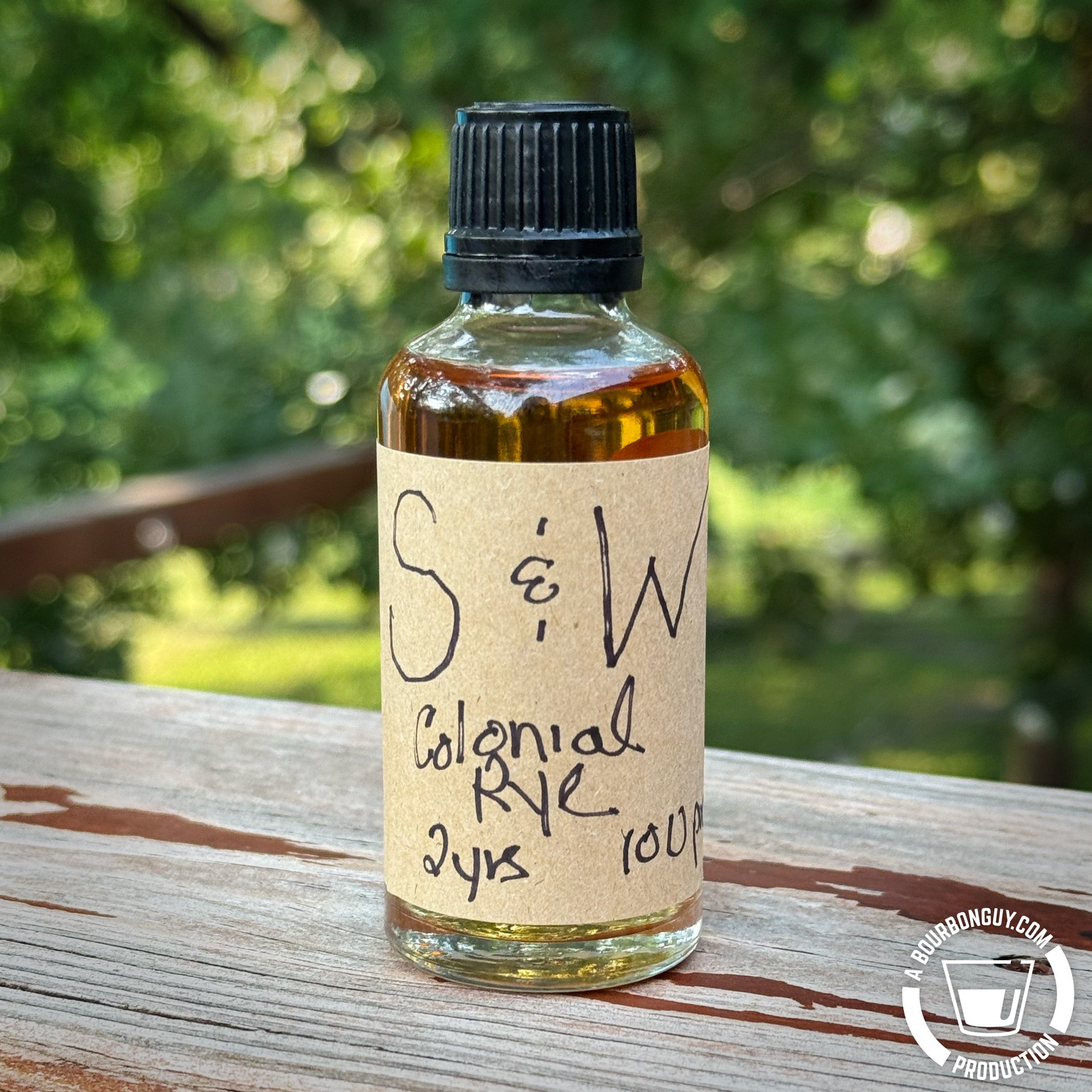 IMAGE: A sample bottle of Wolfe & Wilson Straight Rye, based on a historical recipe. It has a label that says S&W (for Stoll & Wolfe distillery) Colonial Rye. 2 yrs. 100 proof. The bottle sits on a wooden rail with blurred trees in the background.