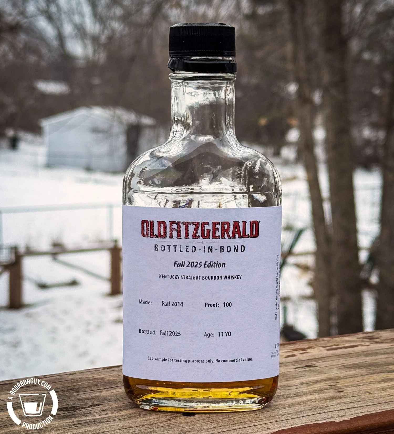 IMAGE: Old Fitzgerald Bottled-in-Bond Fall 2025 Edition bourbon sample bottle on a wooden railing outdoors, snowy backyard and bare trees in the background, showing the minimalist white label and amber whiskey inside.