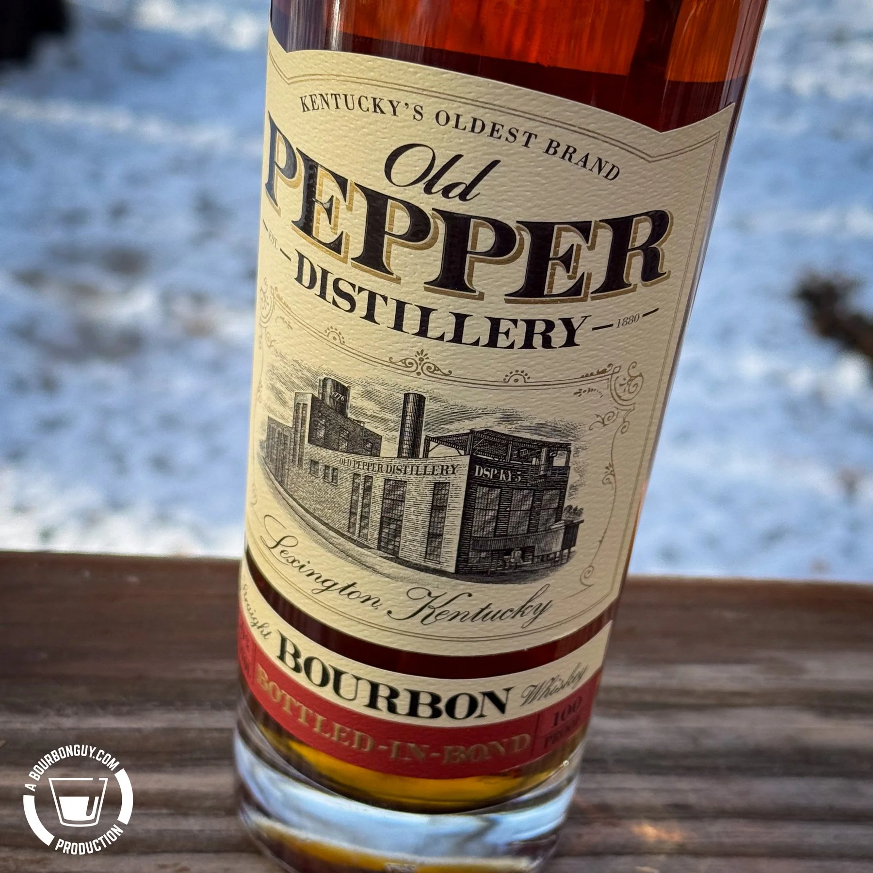 IMAGE: Close-up of an Old Pepper Distillery Bottled-in-Bond Straight Bourbon Whiskey bottle on a wooden railing, with snow-covered ground in the background and the vintage Lexington, Kentucky label in focus.