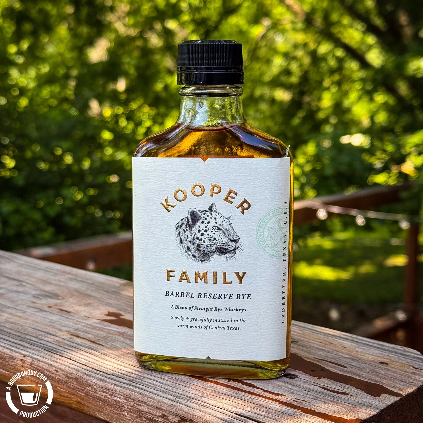 IMAGE: Bottle of Kooper Family Barrel Reserve Rye whiskey standing on a weathered wooden deck railing. The white label features a leopard illustration, with a green, leafy backyard softly blurred in the background.