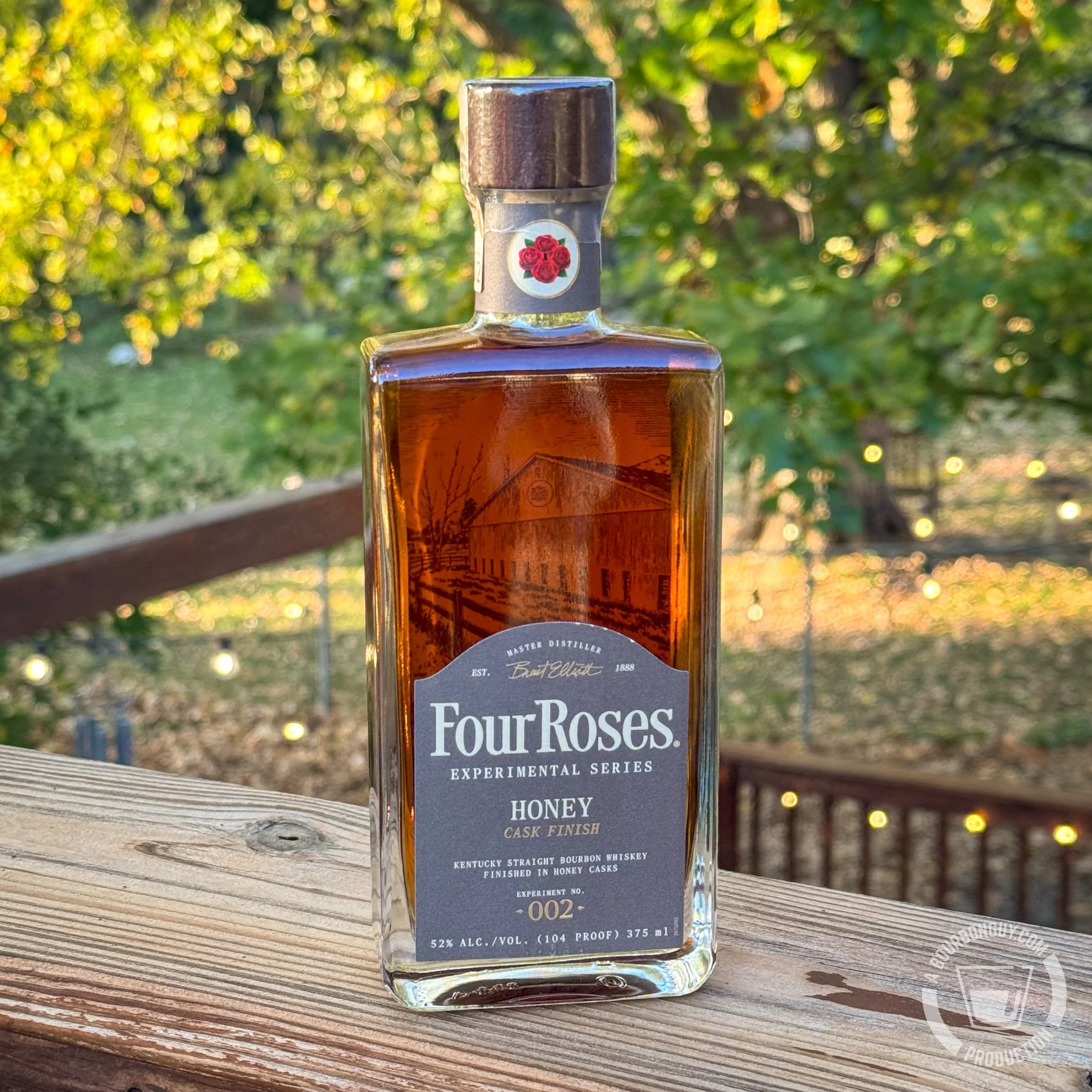 IMAGE: A bottle of Four Roses Experimental Series No. 002 Honey Cask Finish bourbon, 104 proof, sits on a weathered wooden deck railing with sunlit autumn foliage and string lights in the background.