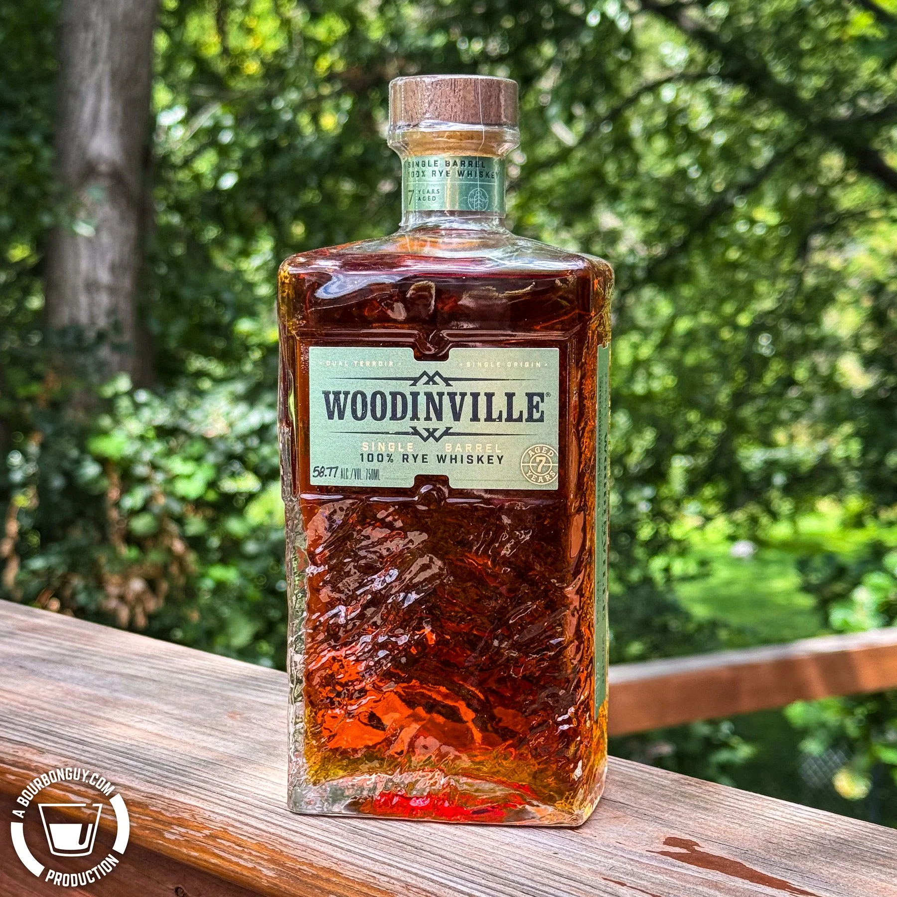 IMAGE: A bottle of Woodinville Single Barrel Cask Strength Rye Whiskey sits on a wooden deck railing against a softly blurred green background.