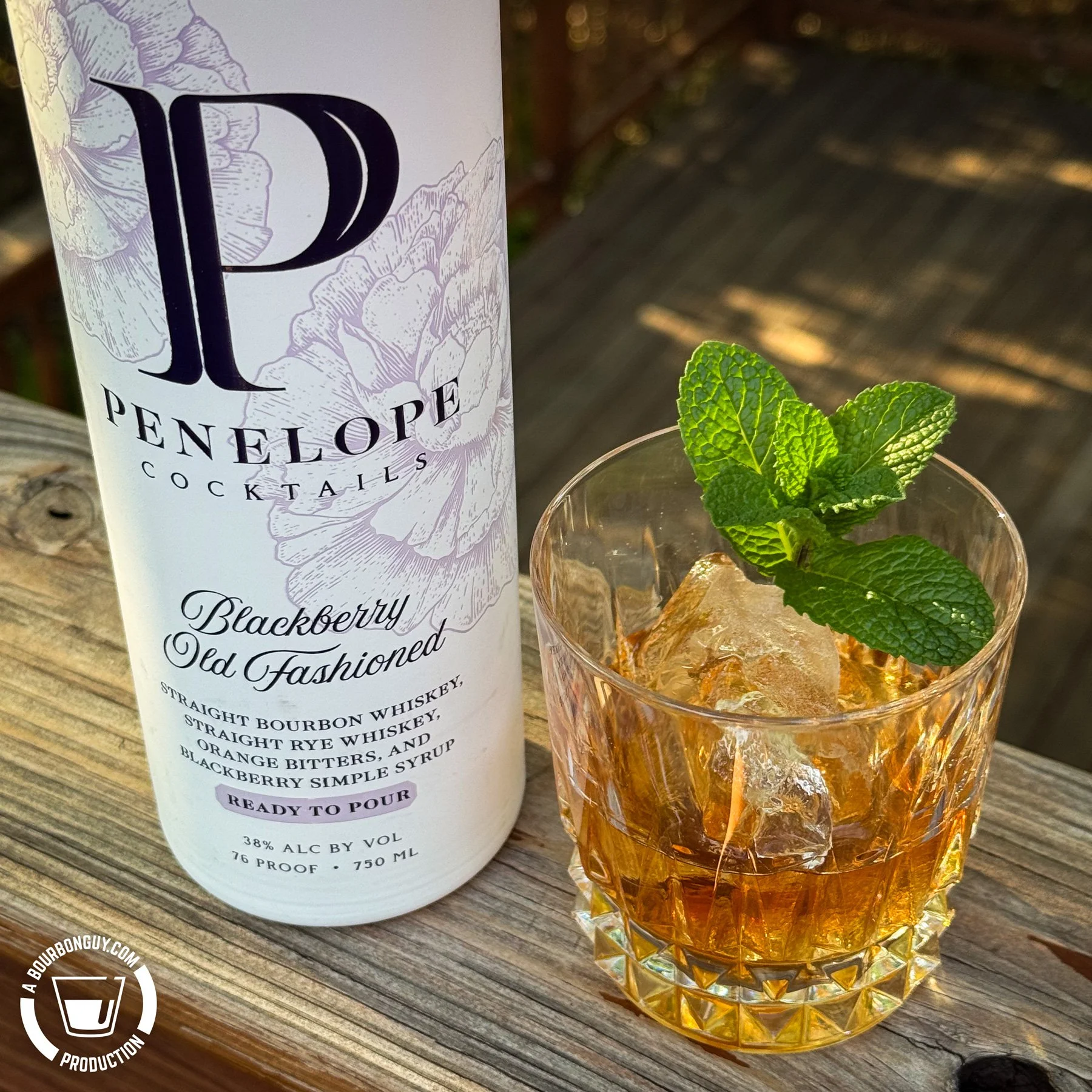 IMAGE: Bottle of Penelope Blackberry Old Fashioned ready-to-pour cocktail beside a rocks glass filled with the drink over ice and garnished with fresh mint. Photographed outdoors on a wooden deck in warm afternoon sunlight.