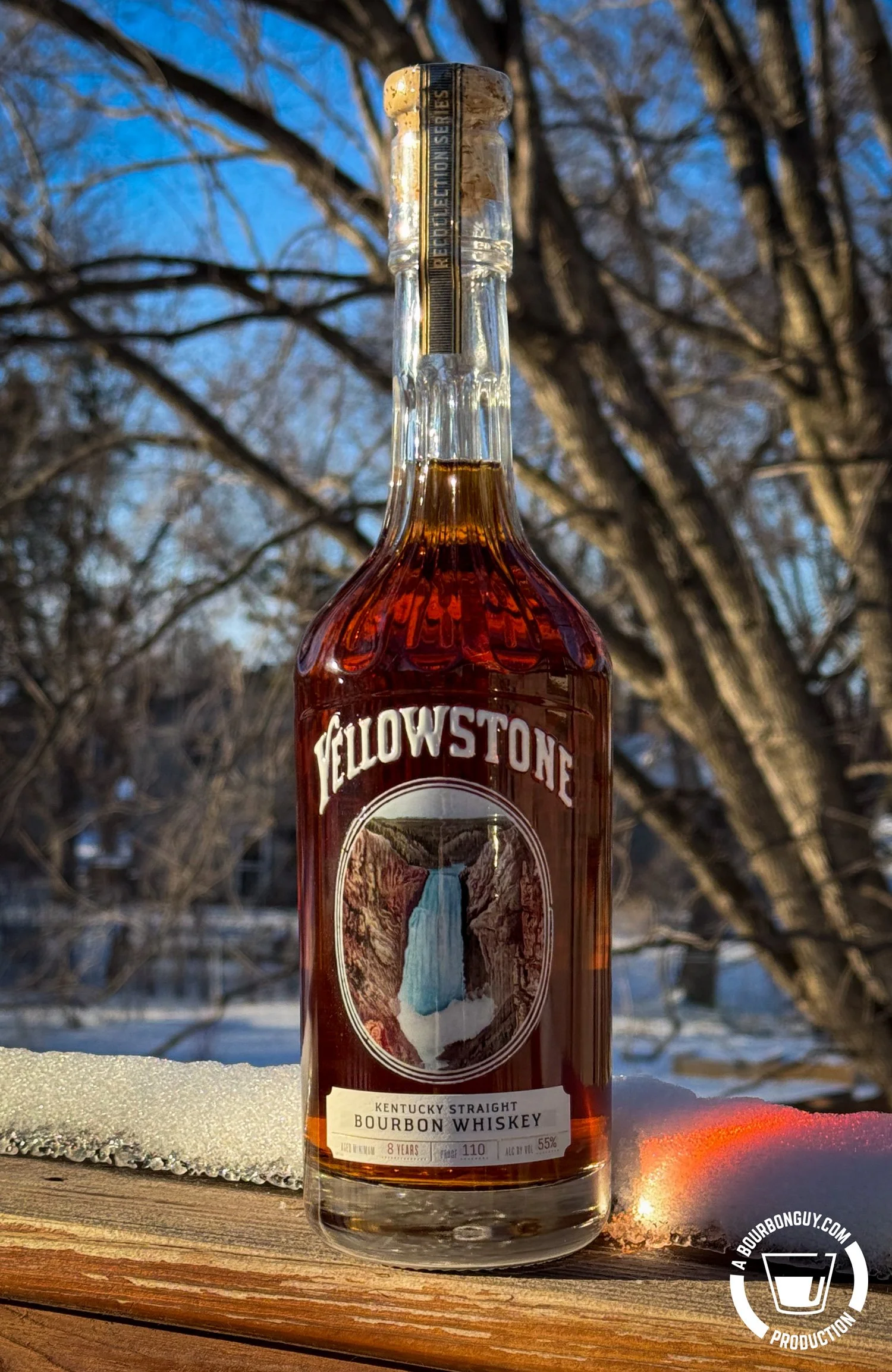 IMAGE: Yellowstone Kentucky Straight Bourbon Whiskey 8 Years, 110 proof, photographed on a snow-covered wooden railing with winter trees in the background and warm sunlight illuminating the amber bottle.