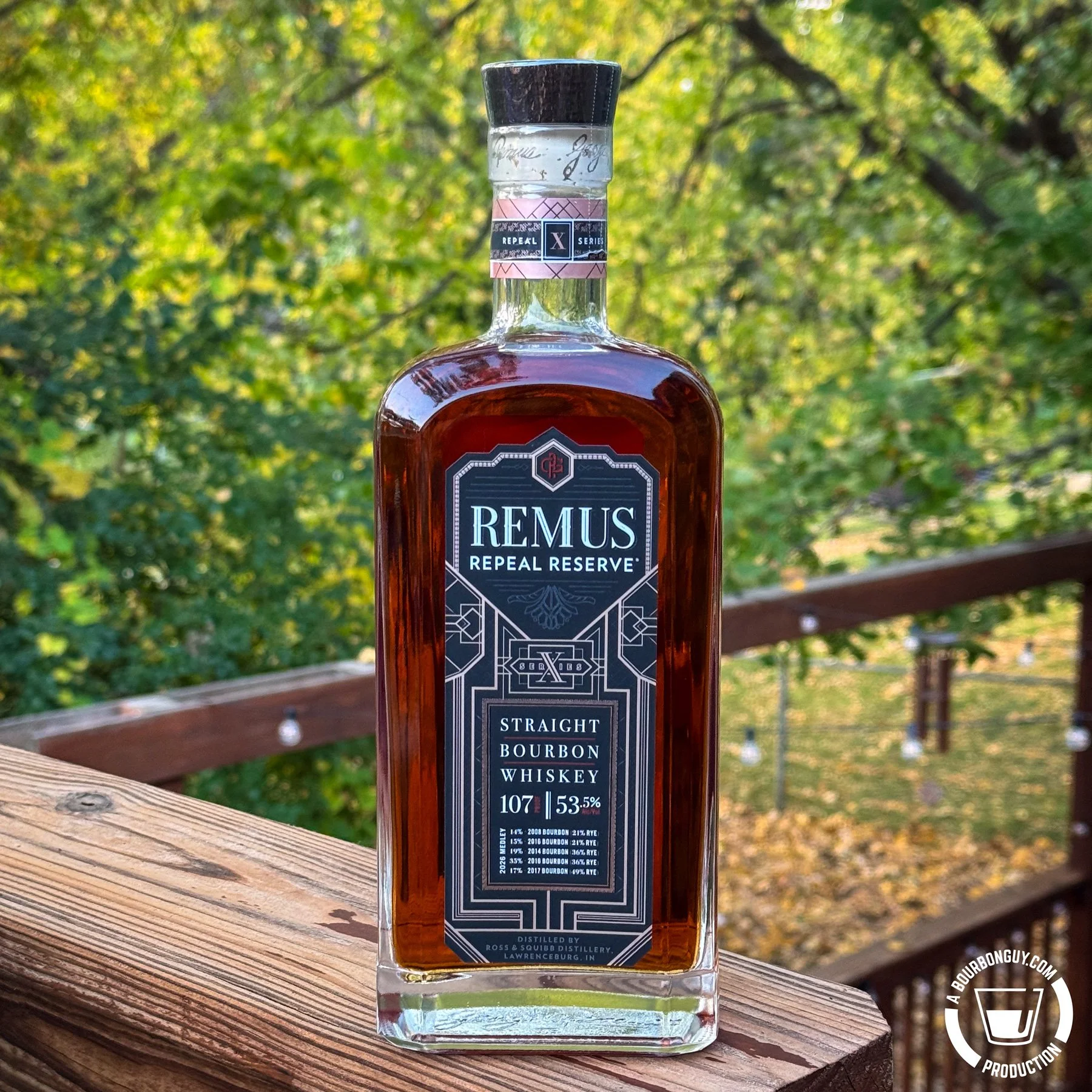 IMAGE: A bottle of Remus Repeal Reserve Series X straight bourbon whiskey, 107 proof, sits on a weathered wooden deck railing with sunlit green and yellow foliage in the background.
