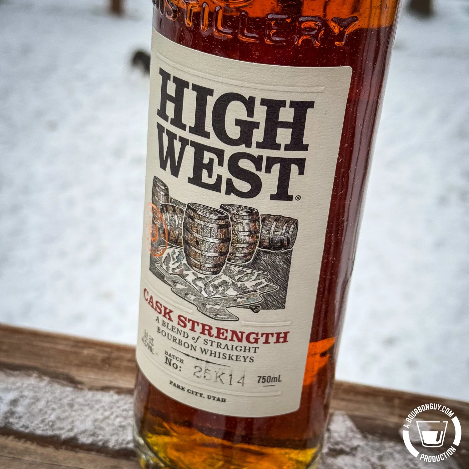 IMAGE: Close-up of a High West Cask Strength bourbon bottle on a snow-dusted wooden railing, showing the label with barrels illustration and amber whiskey, winter background softly blurred.