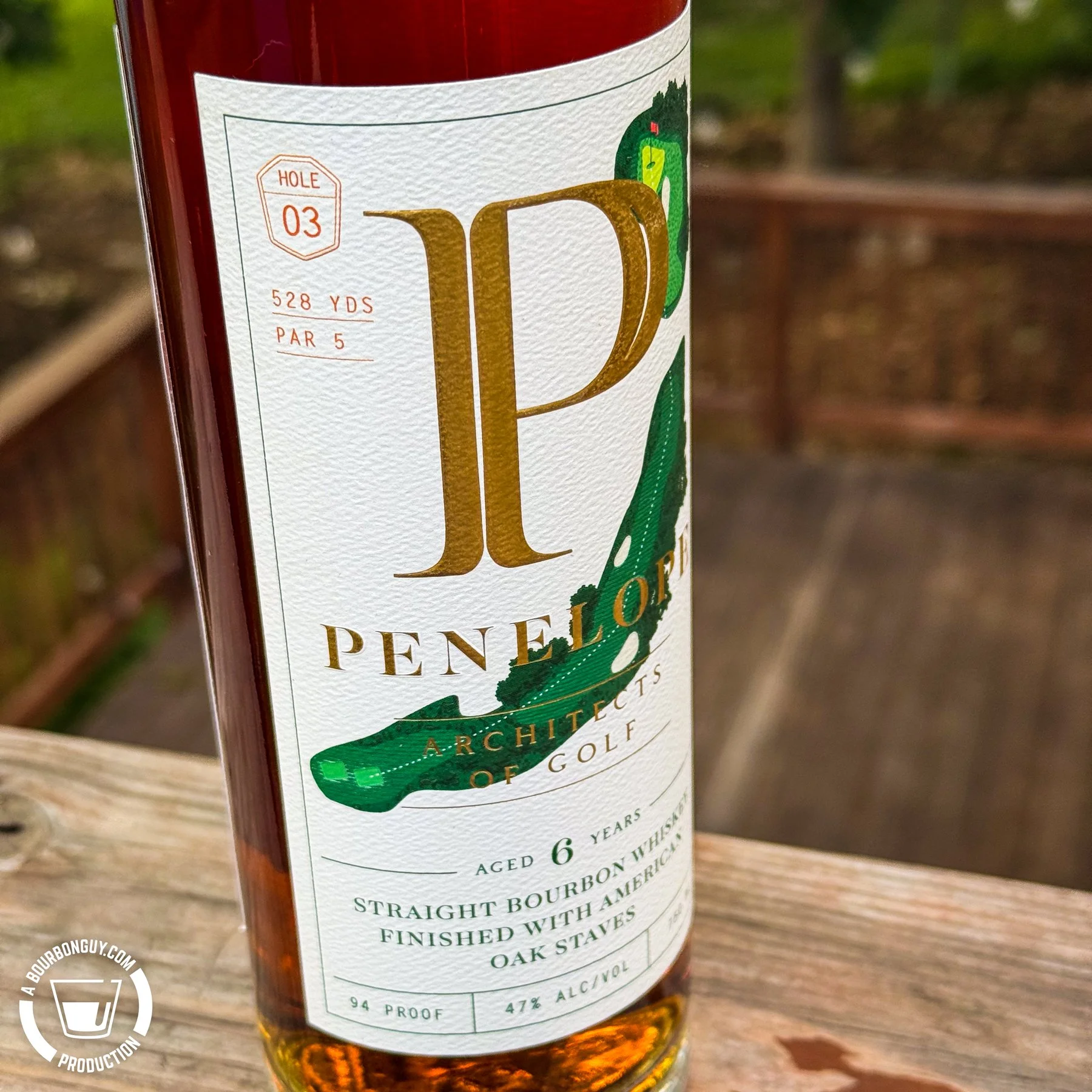 IMAGE: Close-up of a bottle of Penelope Architects of Golf, Hole 3 bourbon on a weathered wooden deck railing. The label features a stylized golf hole design and notes the bourbon is aged six years and finished with American oak staves.