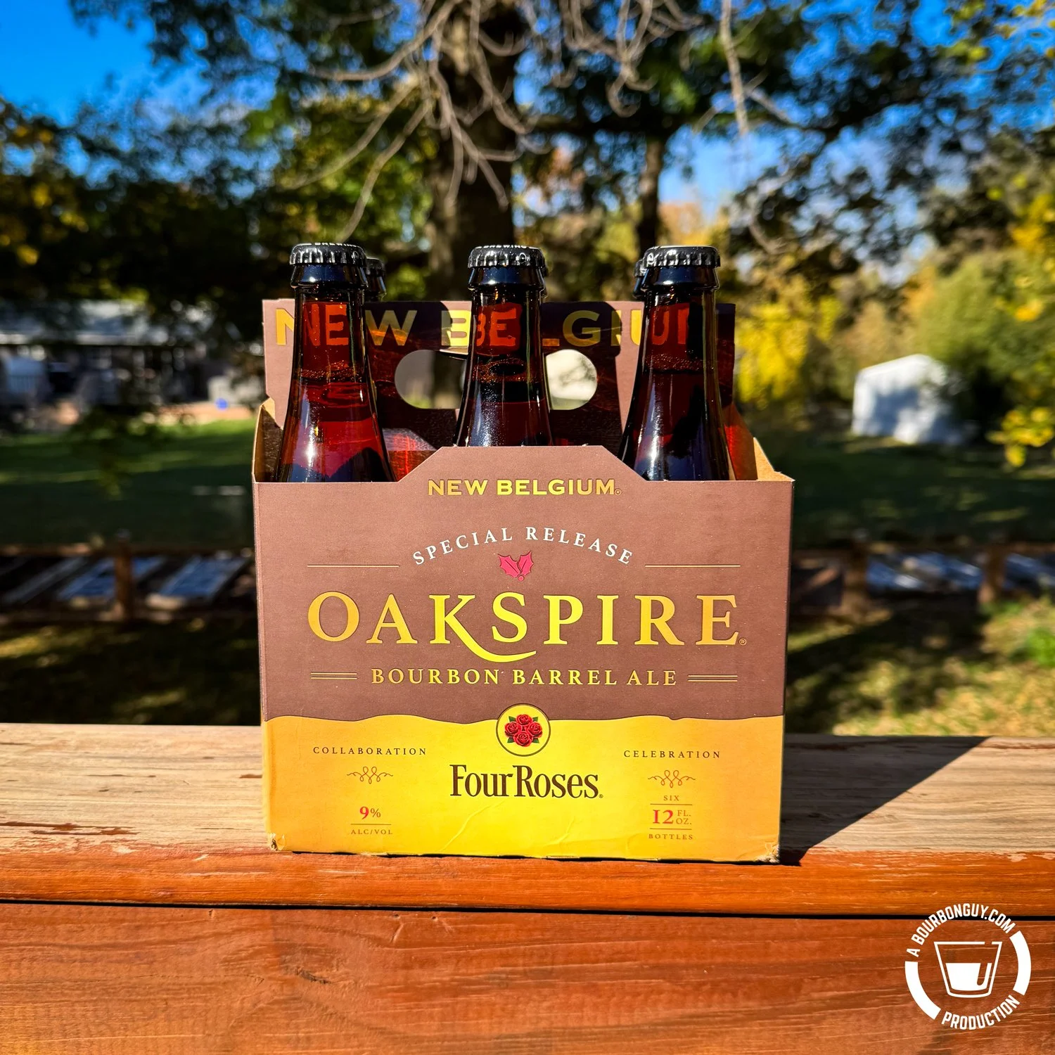 IMAGE: Four-pack of New Belgium Oakspire Bourbon Barrel Ale, a special release collaboration with Four Roses, sitting on a wooden deck rail with green trees and a sunny backyard in the background.