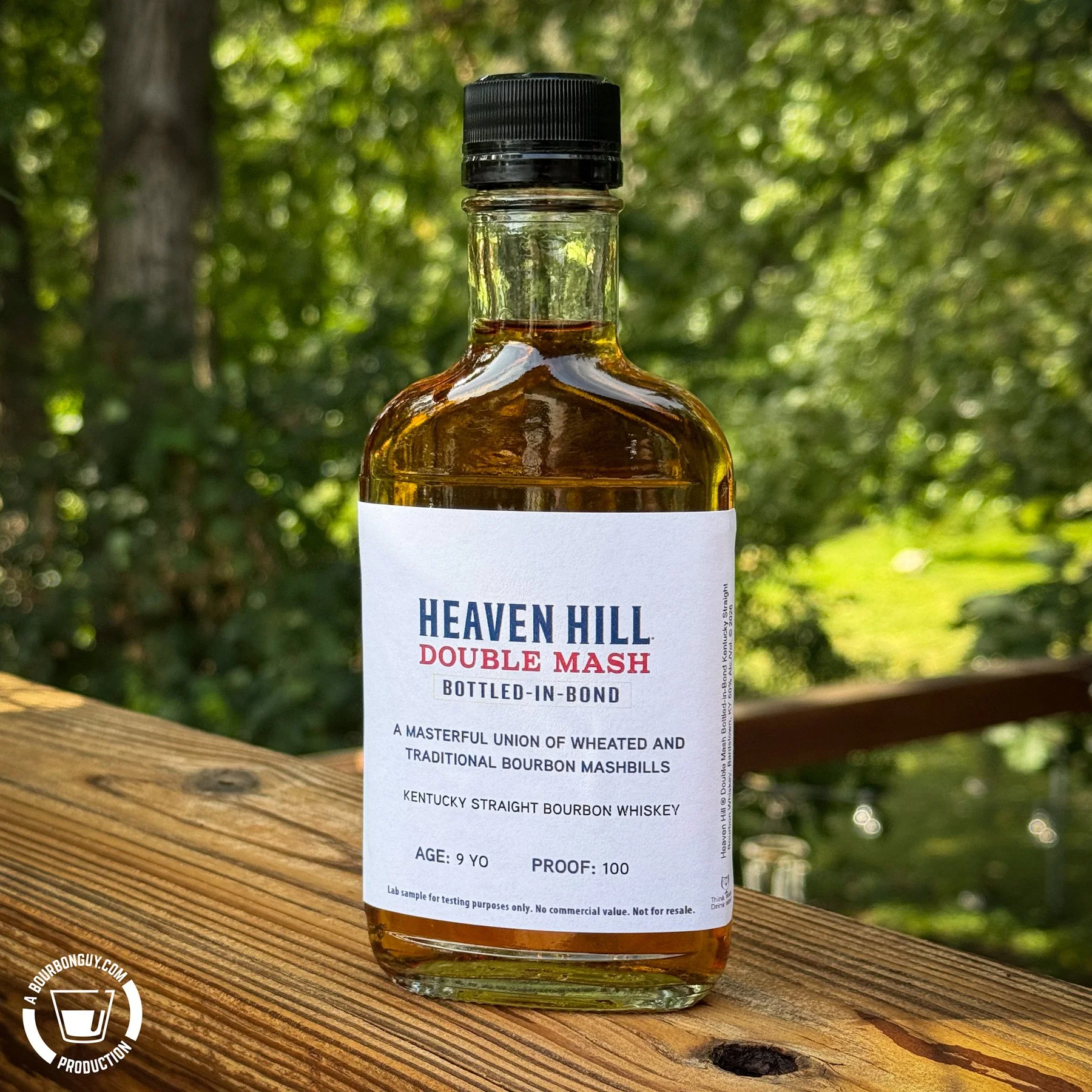 IMAGE: Bottle of Heaven Hill Double Mash Bottled-in-Bond 9-Year-Old Bourbon on a wooden deck railing with a leafy green background.