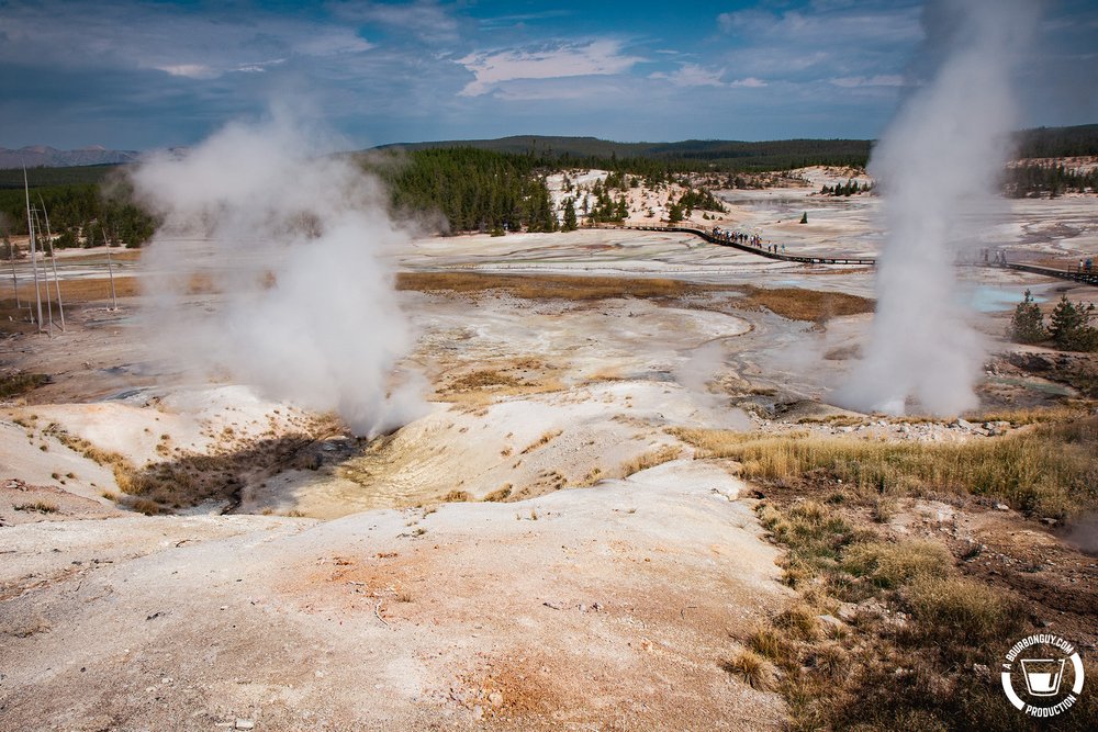 IMAGE: Landscape shot of Yellowstone National Park. Two geysers are in the foreground with tree-covered mountains in the distance.