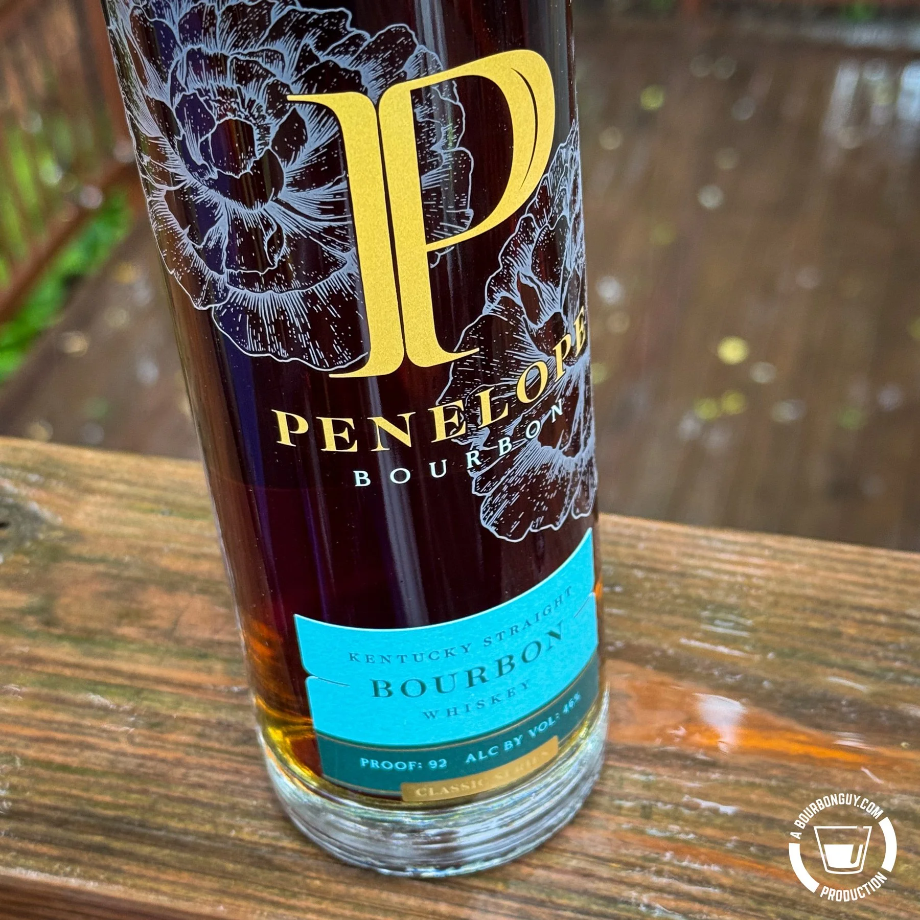 IMAGE: Close-up of a Penelope Straight Bourbon bottle on a rain-soaked wooden deck railing, with the gold logo and blue label prominently displayed against a softly blurred rainy background.