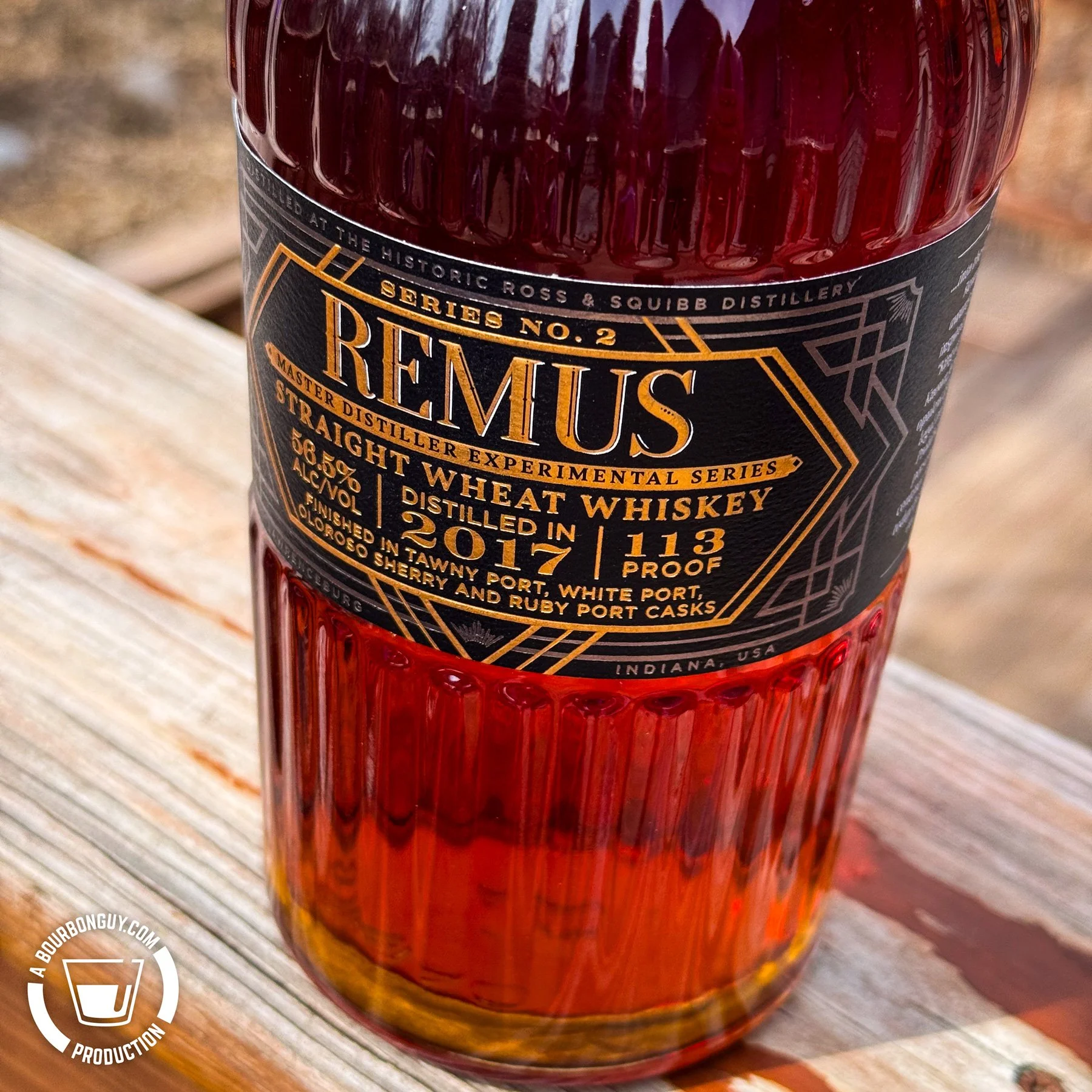 IMAGE: Close-up of Remus Master Distiller Experimental Series No. 2 Straight Wheat Whiskey bottle showing label details (distilled in 2017, 113 proof) on a wooden deck, with rich amber-red whiskey color and BourbonGuy watermark visible