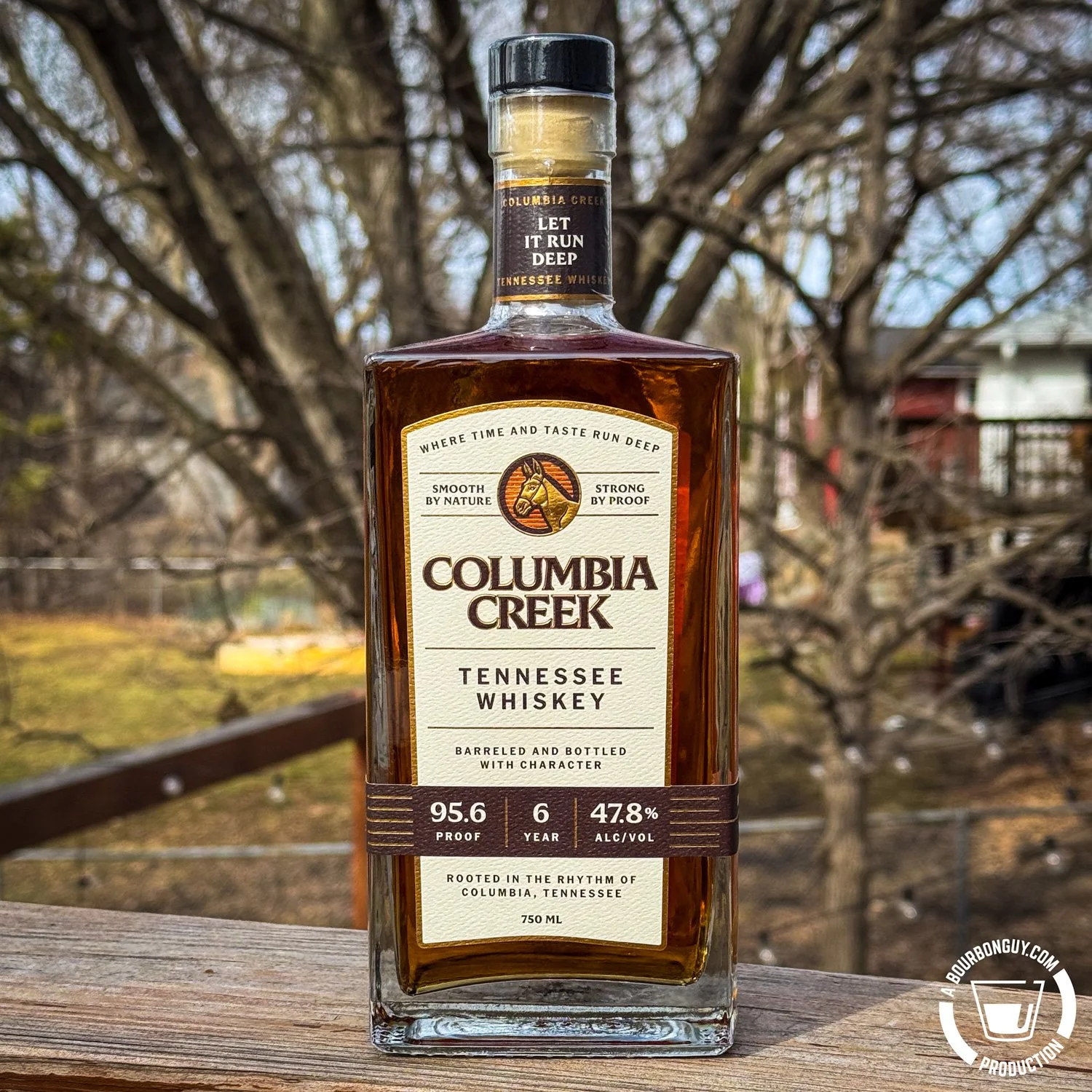 IMAGE: Columbia Creek Tennessee Whiskey bottle, 6-year, 95.6 proof, 47.8% ABV, 750ml, on an outdoor deck railing with bare trees and a red building in the background.