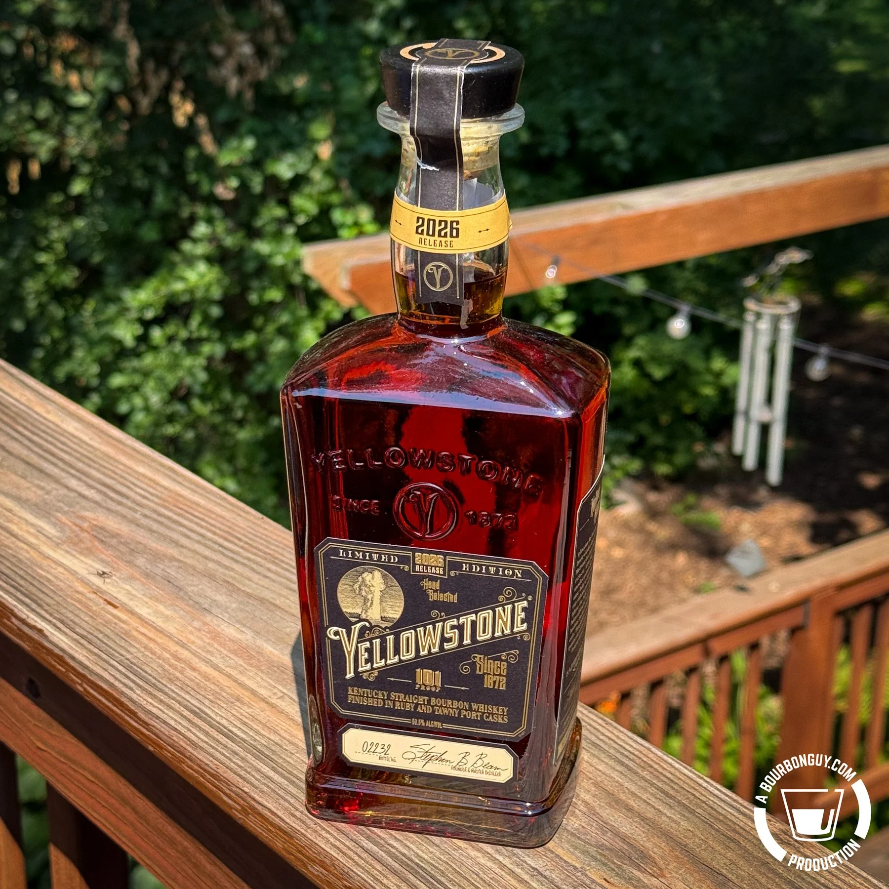 IMAGE: A bottle of Yellowstone Limited Edition 2026 bourbon rests on a wooden deck railing, surrounded by sunlit green foliage.