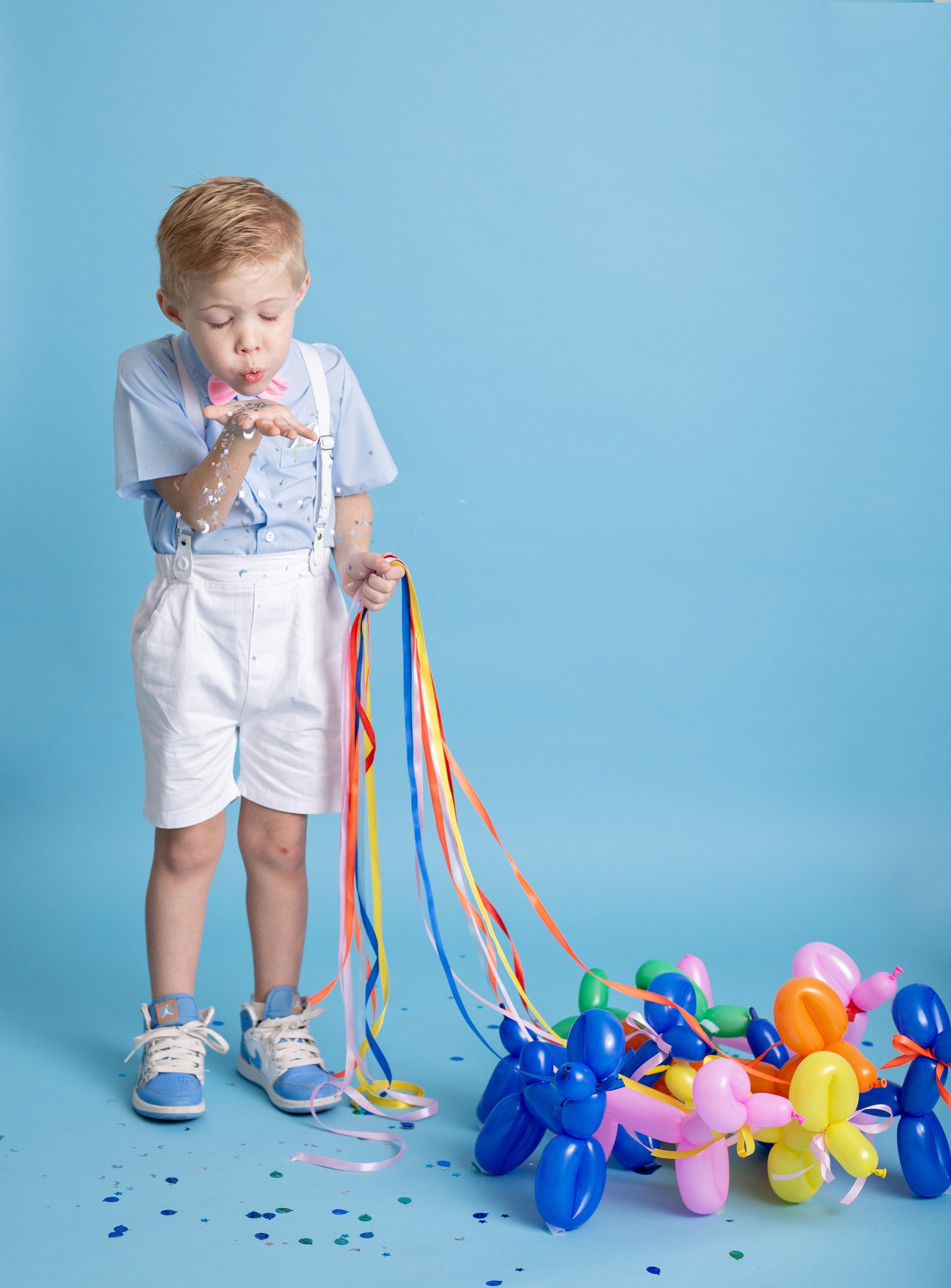 A happy young boy smiles during a colorful birthday photo session in Gilbert, Arizona, wearing suspenders and a bow tie while holding balloon dog decorations against a bright blue backdrop.