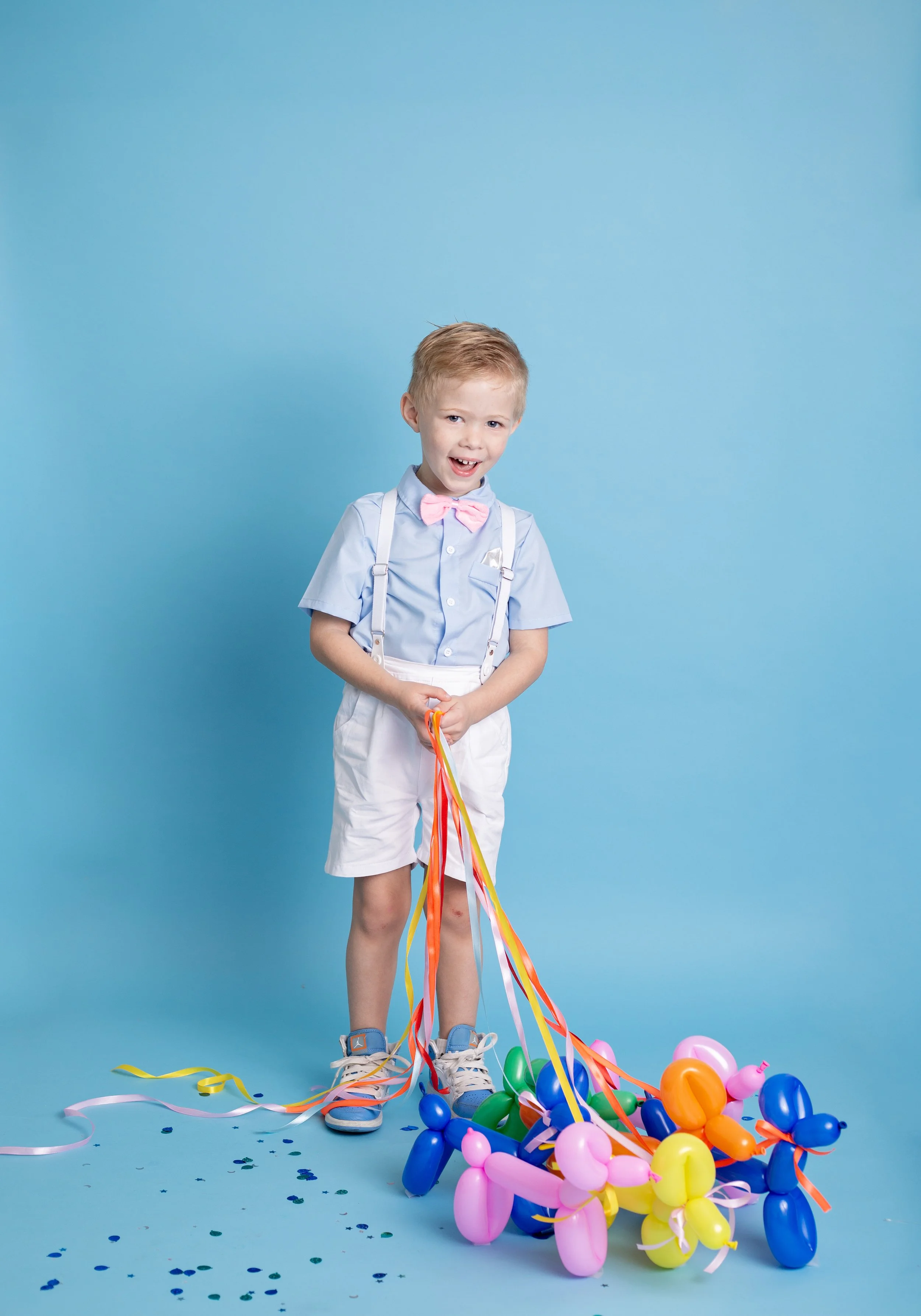 A happy young boy smiles during a colorful birthday photo session in Gilbert, Arizona, wearing suspenders and a bow tie while holding balloon dog decorations against a bright blue backdrop.