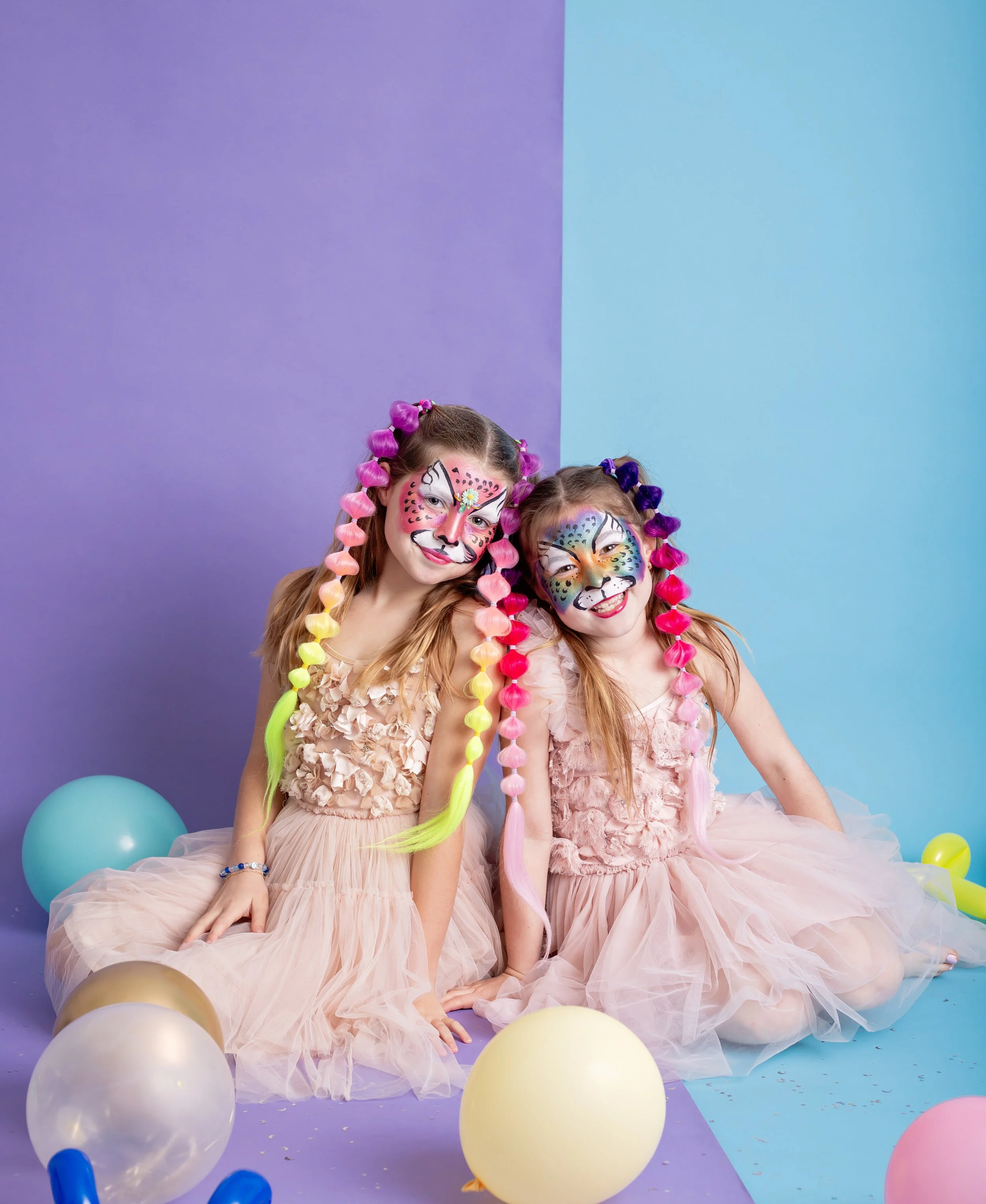 young girls with colorful braided hair extensions and vibrant animal face paint sit together during a studio branding session for an East Valley Arizona face painter, photographed by a Gilbert Arizona child photographer.