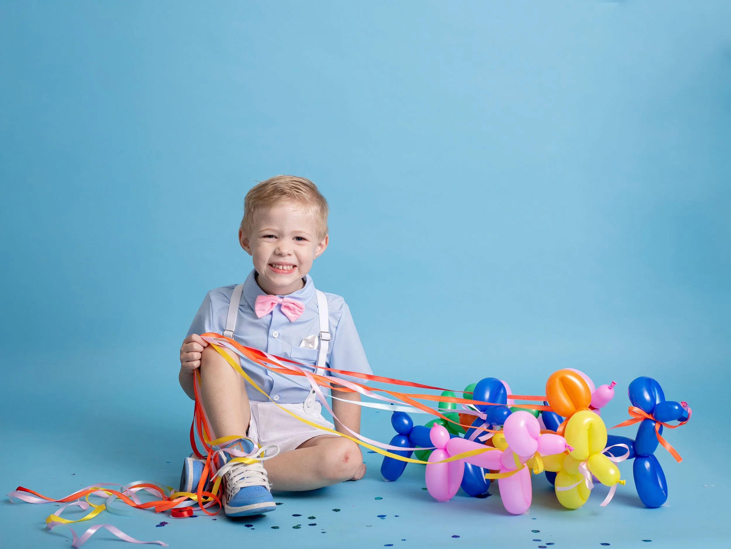 A happy young boy smiles during a colorful birthday photo session in Gilbert, Arizona, wearing suspenders and a bow tie while holding balloon dog decorations against a bright blue backdrop.
