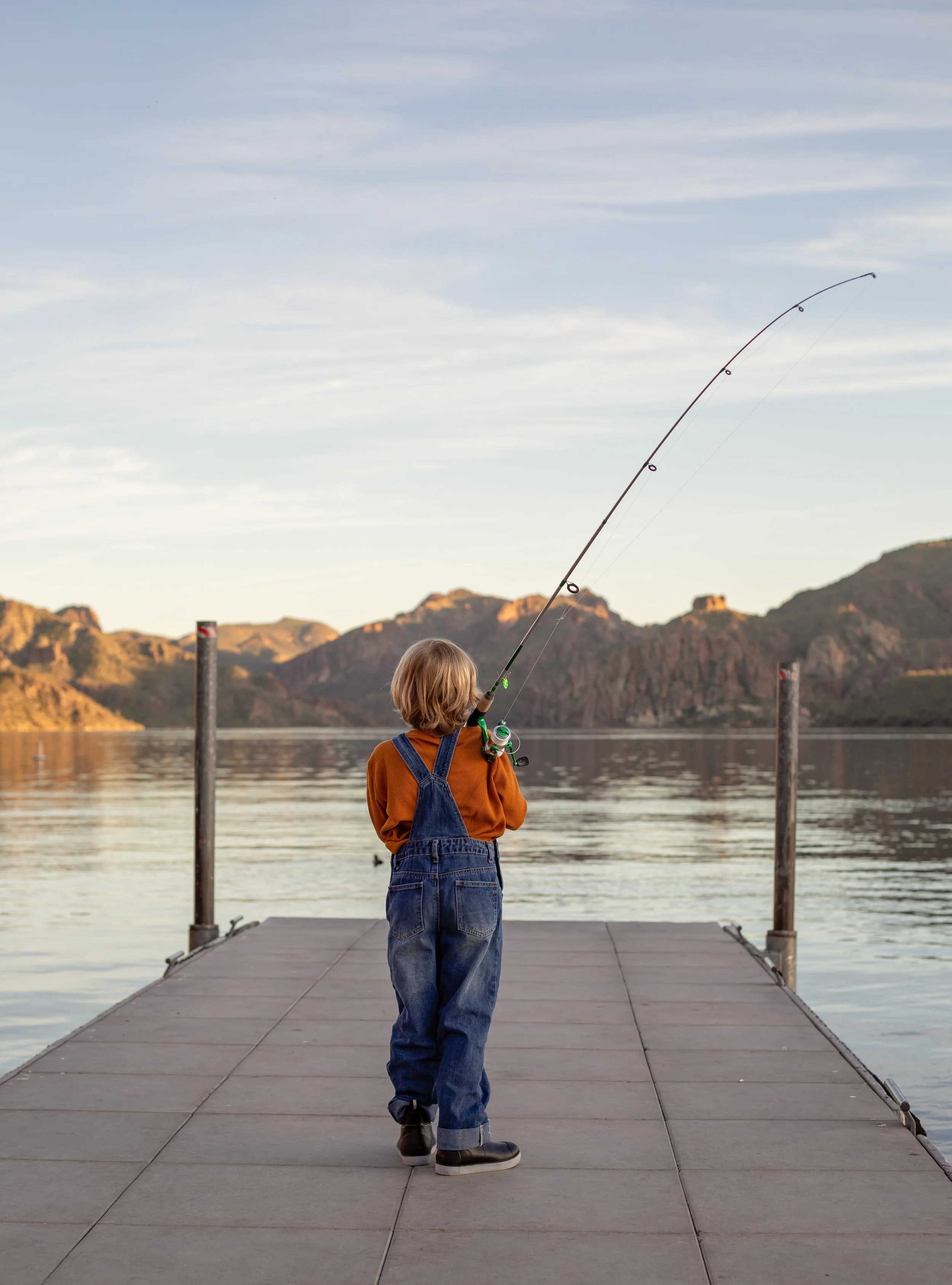 Child sitting on a dock holding a fishing pole and watching the water at sunset in Arizona, photographed by a Gilbert family photographer