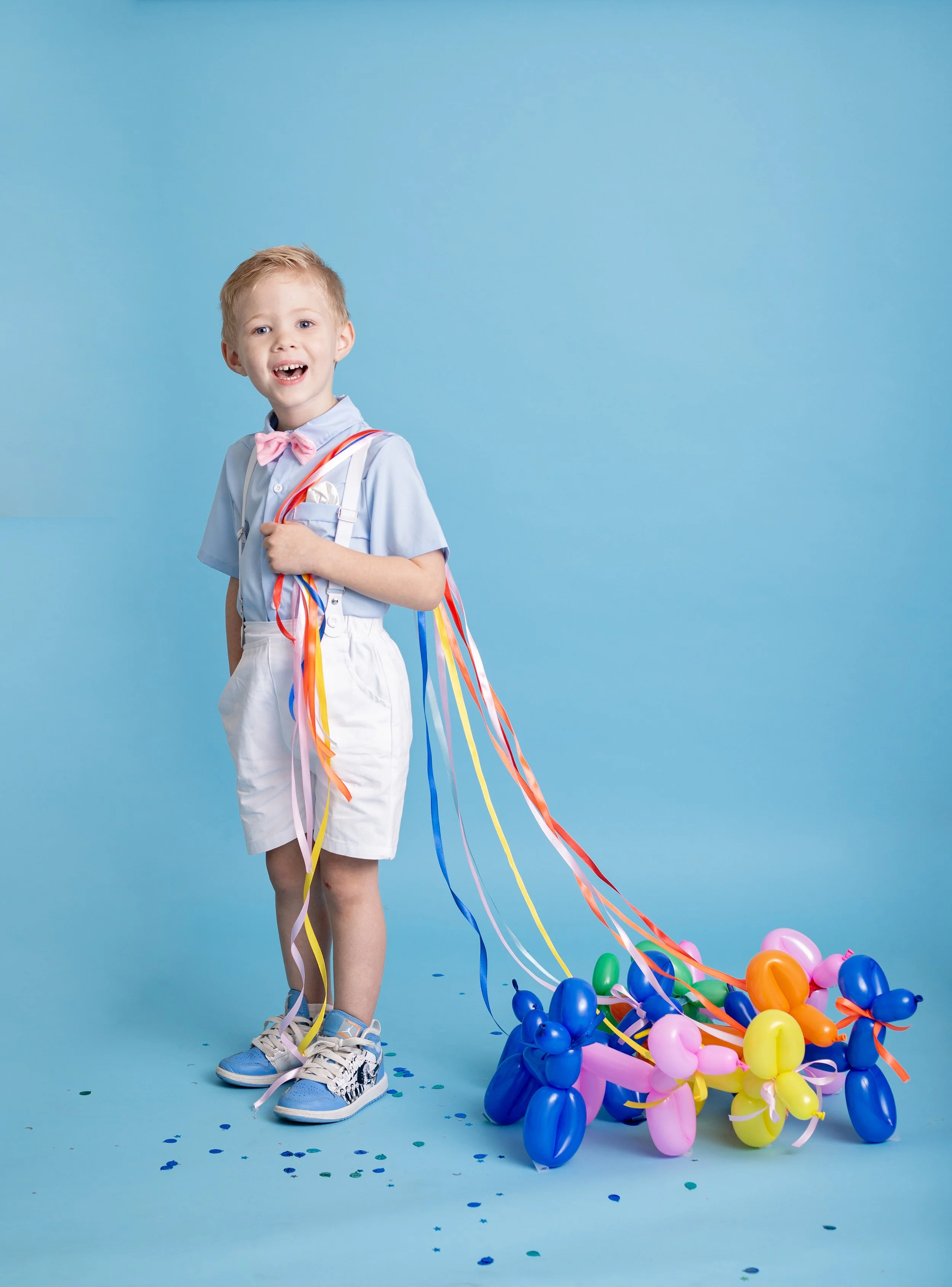 A happy young boy smiles during a colorful birthday photo session in Gilbert, Arizona, wearing suspenders and a bow tie while holding balloon dog decorations against a bright blue backdrop.