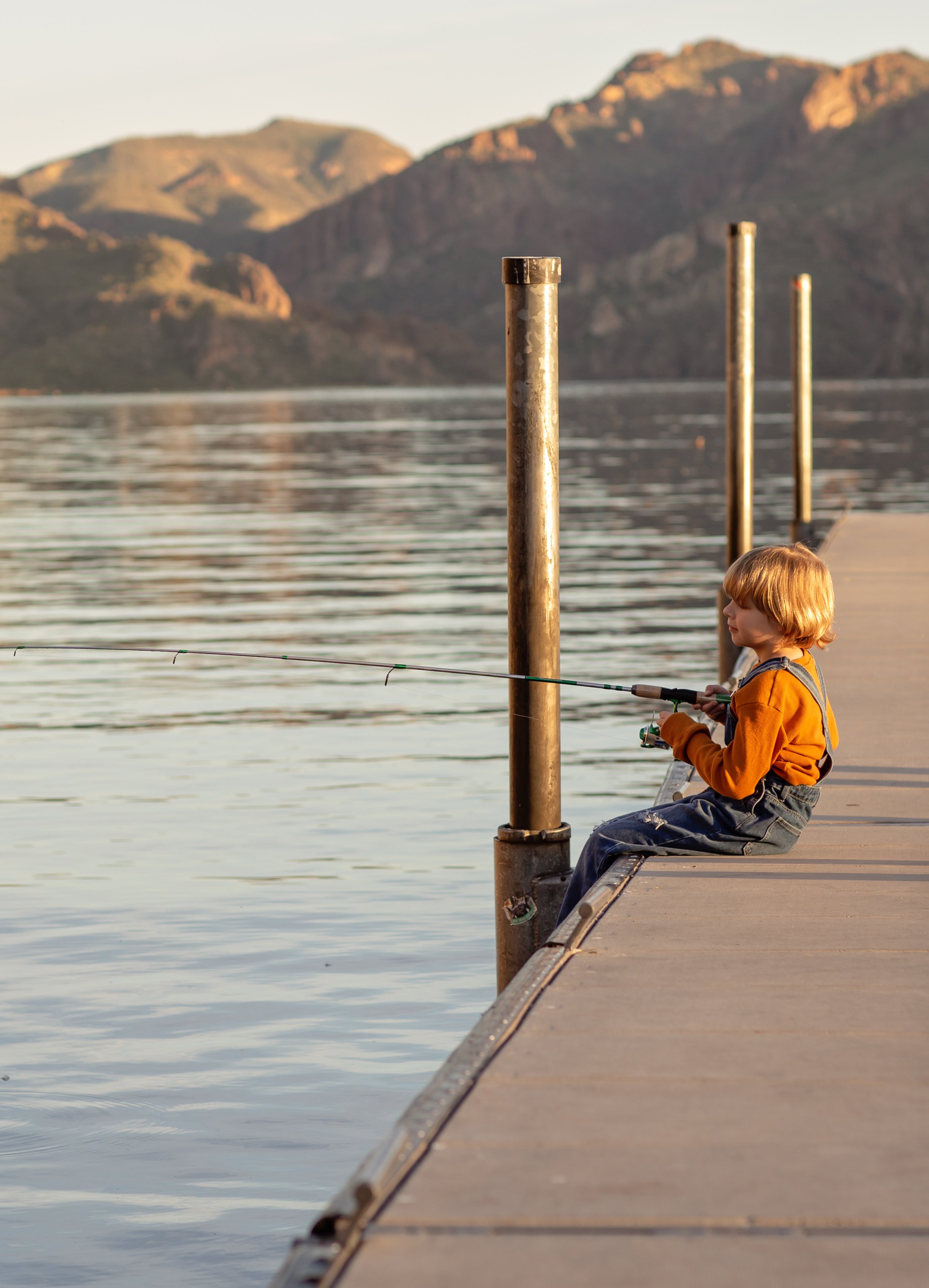 Child sitting on a dock holding a fishing pole and watching the water at sunset in Arizona, photographed by a Gilbert family photographer